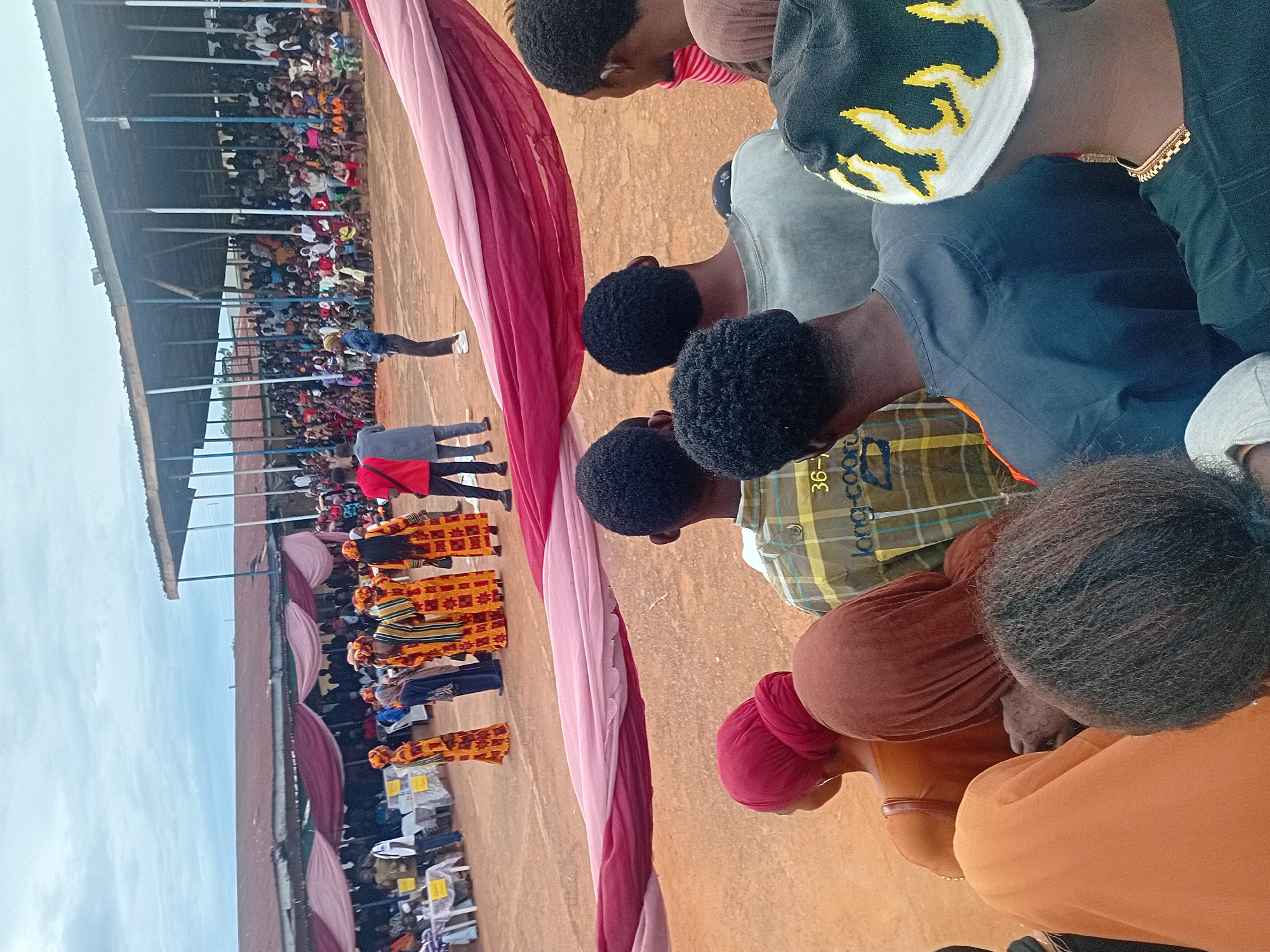 The image captures an outdoor public event in Ogbogbo, Nigeria, under an overcast sky, viewed from behind a cluster of onlookers.
In the immediate foreground, the backs of approximately ten individuals are visible. These include several males with dark, tightly curled hair; one wears a black beanie with a yellow and white flame design, and another is clad in a green and yellow plaid shirt with "long-coo" text visible. Multiple females are also present, distinguished by head coverings in solid colors such as red, brown, and orange.
Stretching horizontally across the mid-foreground is a large fabric, faded pink merging into dark red, acting as a decorative element or a soft barrier. Beyond this fabric, the central event area is visible on a reddish-brown dirt ground. Here, a group of at least six individuals, likely performers, are positioned. They are dressed in striking traditional attire featuring bright yellow, orange, and black patterns, standing in a formation. Near the performers, two men observe: one in a red shirt and dark trousers, and another in a light grey tunic over dark trousers. Further left, a man in a blue shirt stands near a section with several white-covered tables marked with yellow placards, indicating a designated area.
The background features a large, crowded grandstand filled with numerous spectators of varying ages, all observing the event. The grandstand is characterized by a corrugated metal roof supported by a series of vertical blue and white poles. Beyond the grandstand, additional low-rise buildings with reddish-brown roofs are present under the uniformly cloudy sky.