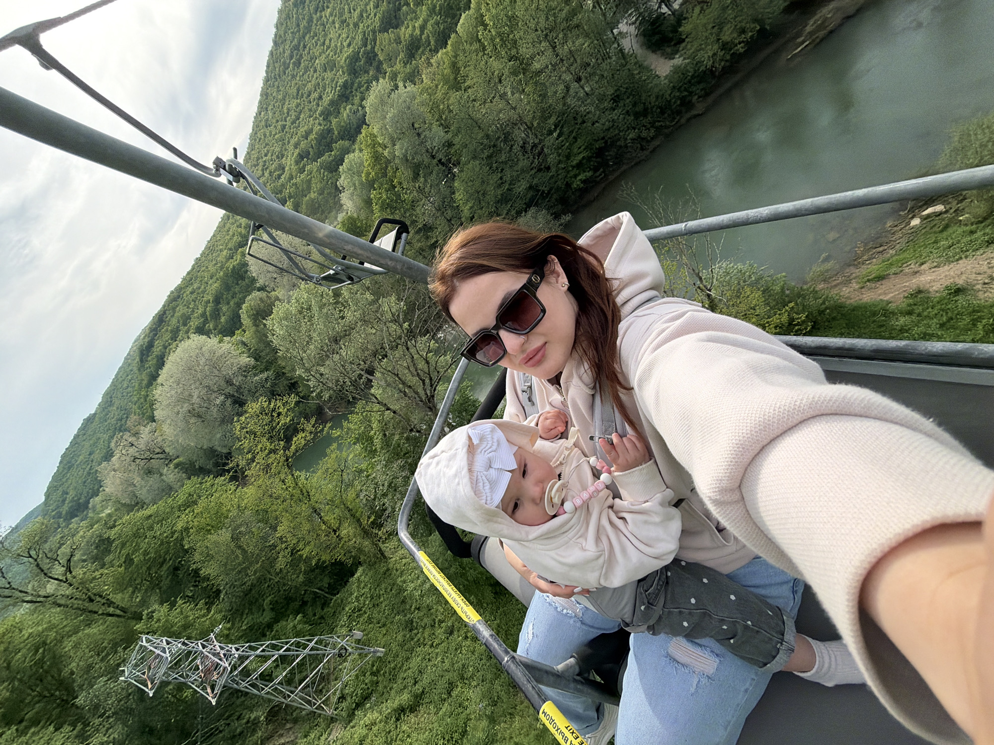 A selfie captures a woman and baby riding a chairlift high above the city of Goryachiy Klyuch, Russia. The woman, wearing sunglasses and a light-colored hooded sweatshirt, holds the baby in her lap. The baby is wearing a hooded outfit and has a pacifier. The chairlift ascends over a lush green landscape, with trees covering the hillsides. A river can be seen winding through the valley below. The gray metal infrastructure of the chairlift is visible in the foreground.