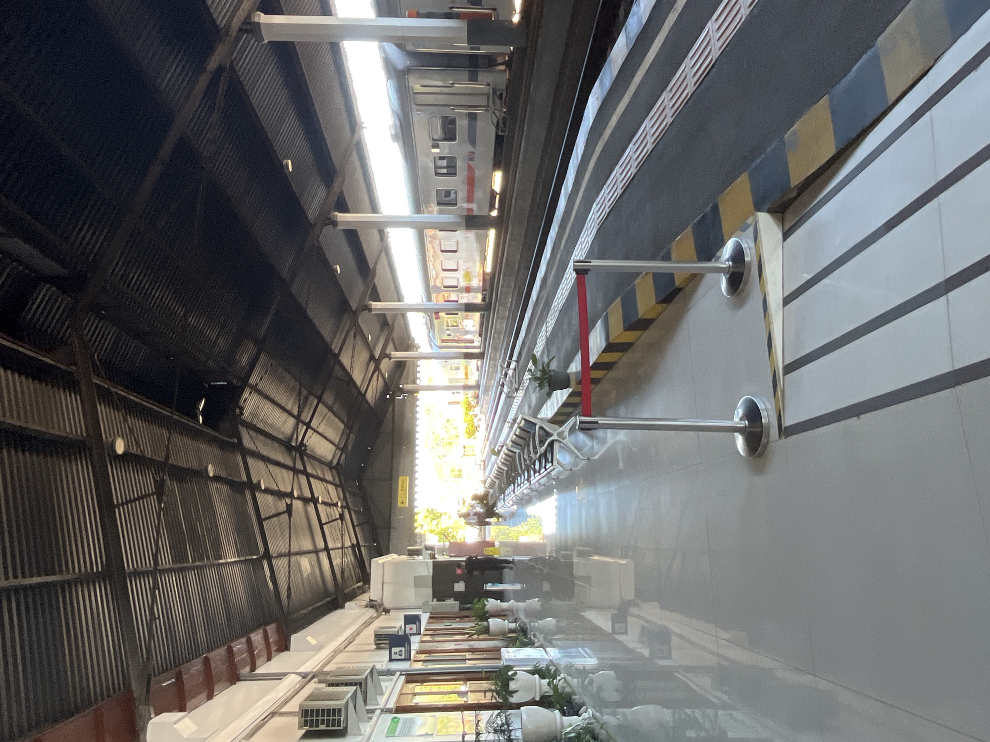 The image shows an outdoor view of a train station platform in Desa Tambakreja, Indonesia. The platform is covered by a dark, corrugated metal roof supported by white pillars. To the left, a white building has several windows and air conditioning units mounted on the exterior. Potted plants are placed along the walkway. The walkway itself is paved with light-colored tiles with reflective surfaces. Several rows of empty white chairs are lined up on the platform. A red rope barrier with stanchions is positioned along the edge of the platform. The train tracks are visible to the right, with a silver train parked alongside. Yellow and black checkered patterns mark the edge of the platform. In the distance, there are glimpses of trees and sunlight. Two people are visible in the background, one walking and others sitting.