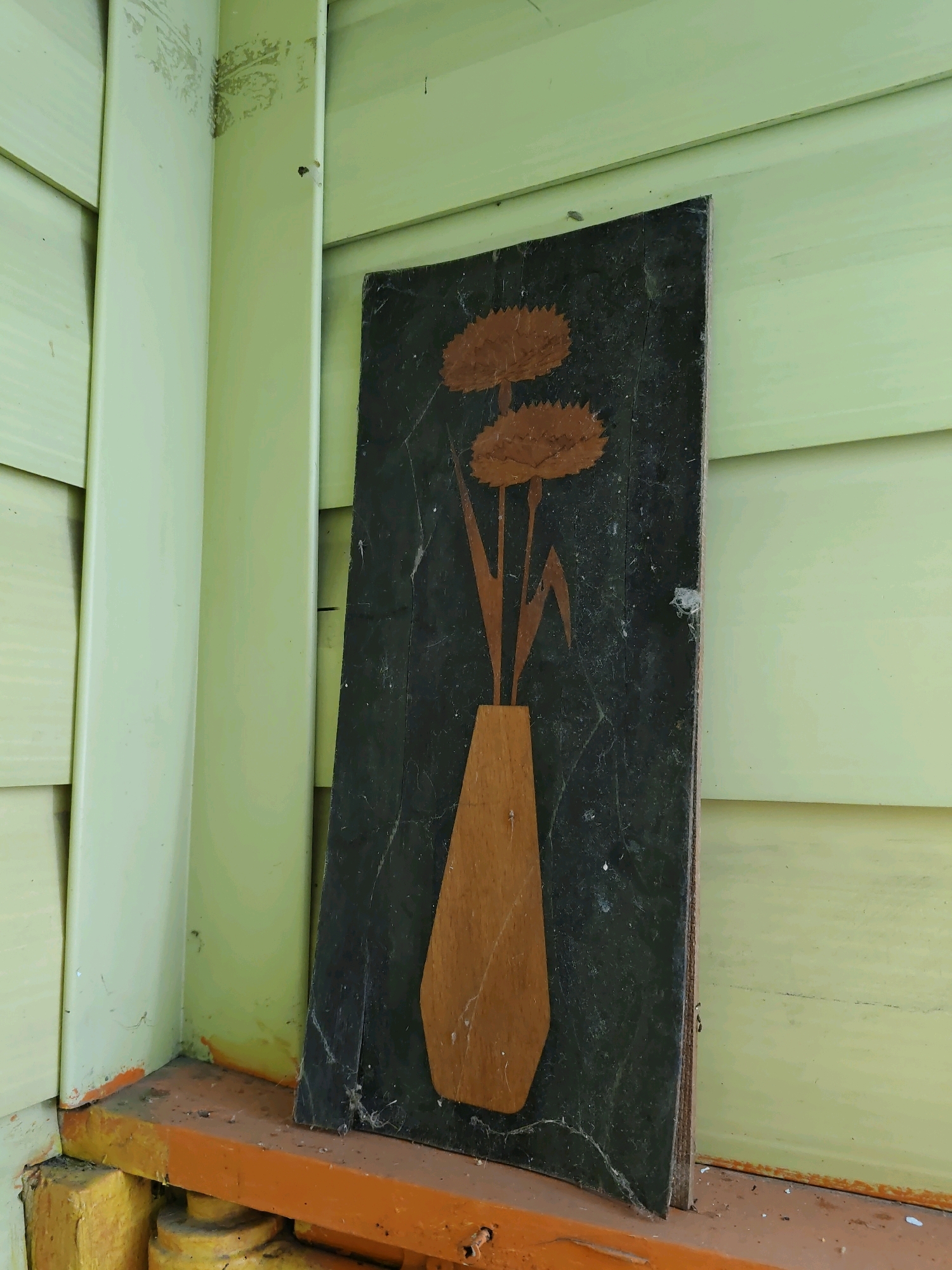 A vertical, dark-colored panel featuring a wooden inlay of a vase with two flowers stands propped against a pale green, clapboard-sided wall, likely outdoors. The panel is positioned in a corner, leaning on a weathered, orange-painted ledge. The surrounding wall exhibits signs of age and weathering, with some peeling paint and minor stains near the base. The overall scene suggests an outdoor storage area or a neglected exterior corner of a building.