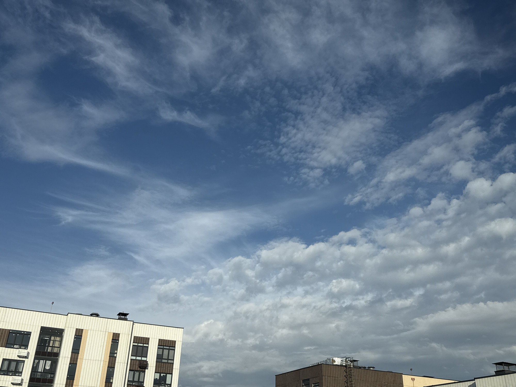 The image captures a view of the sky above buildings in Lyubertsy, Russia, on a day with a dynamic cloudscape. The sky is a vibrant blue, transitioning to lighter shades near the horizon, and is filled with a variety of clouds. Wispy cirrus clouds streak across the upper portion of the sky, while denser, more textured cumulus clouds populate the lower half.
In the foreground, the rooftops and upper facades of several multi-story buildings are visible. The building on the left is modern, with a light-colored, tiled exterior and numerous windows. Some air conditioning units are mounted on the exterior walls. The building on the right appears to be older, with a darker brick facade and visible ventilation or structural elements on its roof. A smaller, light-colored building is partially visible in the lower right corner. No people are discernible in the image, and no specific actions or interactions are depicted. The scene suggests an urban environment under a partly cloudy sky.