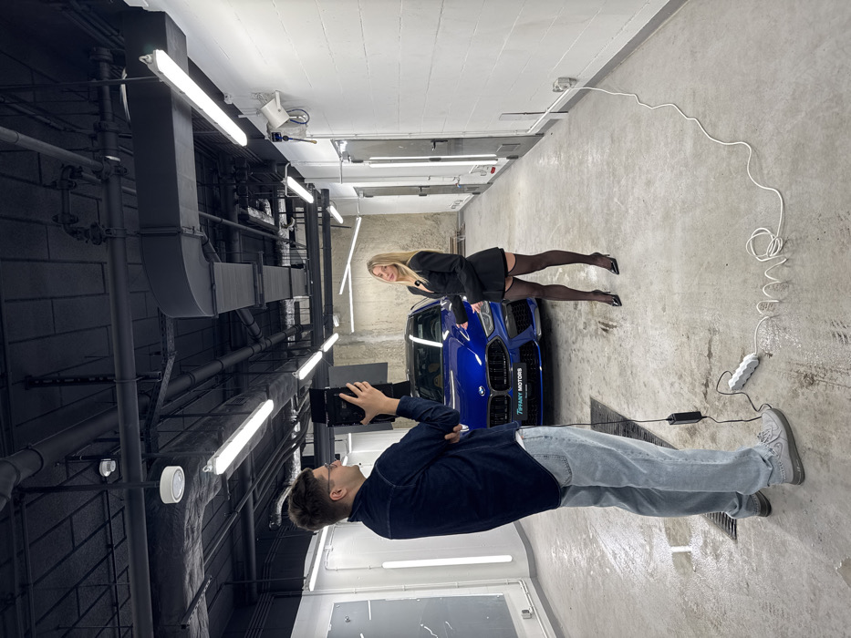 An indoor scene depicts a video production at a Tiffany Motors facility in Warsaw, Poland, identifiable by the "TIFFANY MOTORS WA" license plate on the vehicle. A male individual, appearing to be in his late 20s to early 30s, stands facing right, holding a black professional video camera towards a female subject and a car. He wears a dark blue long-sleeved shirt, light blue jeans, and grey sneakers.
A female individual, in her 20s or early 30s with long blonde hair, stands to the right, facing the camera. She wears a black blazer, black shorts, black sheer stockings with visible garter straps, and black pointed high heels. A small black lapel microphone is clipped to her blazer. She extends her left hand in a gesturing motion towards a metallic blue BMW M8 Coupe positioned between the two individuals. The car is highly polished, with its front grille and headlights prominent.
The environment is a brightly lit, industrial-style space, likely a car detailing studio or showroom. The floor is light grey concrete, with a drain cover visible on the left. The walls are white, with vertical LED light strips on the left and two grey metal doors and a white industrial speaker mounted on the right. The back wall behind the car is a rough, textured grey concrete with horizontal LED light strips. The ceiling is black, featuring exposed ductwork, pipes, and numerous fluorescent and LED lighting fixtures. A white multi-outlet power strip and a black power adapter are on the floor near the male's feet, with a white electrical cord extending to the right. The scene depicts a commercial shoot for the luxury vehicle.