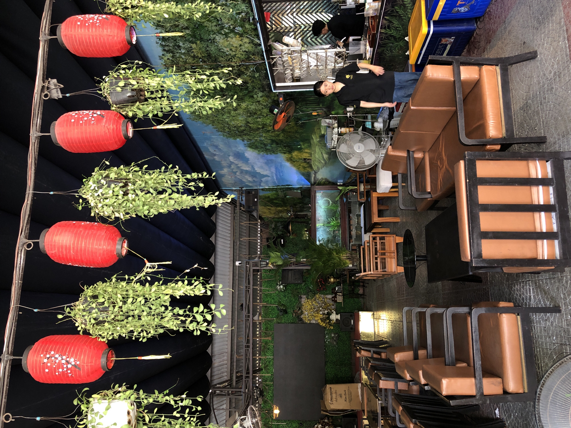 The image shows an outdoor seating area of a cafe or restaurant in Da Nang, Vietnam. The setting is decorated with red paper lanterns and hanging plants, suggesting a vibrant and lively atmosphere.
In the foreground, there are several rows of brown leather booth-style seating with dark metal frames. In the background, there are tables and chairs, some made of wood and others with dark tabletops. A large mural of a forest and mountains covers the back wall, creating a sense of depth and natural beauty.
There are a few people visible in the scene. One person, wearing a black uniform, is standing by a counter, possibly a server or staff member. Another person, also in dark clothing, is working behind the counter. A third person, in a black t-shirt and jeans, is standing near the seating area and looking towards the camera.
A large fan is positioned in the lower left corner of the image, and another fan is visible in the background. There are also several coolers, one blue and yellow, stacked near a counter. A cardboard box with visible text is placed on the floor near some of the seating.
The image appears to be taken during the daytime, with ample natural light suggesting it's not nighttime. The overall impression is a casual and welcoming establishment, possibly a popular spot for locals and tourists in Da Nang.