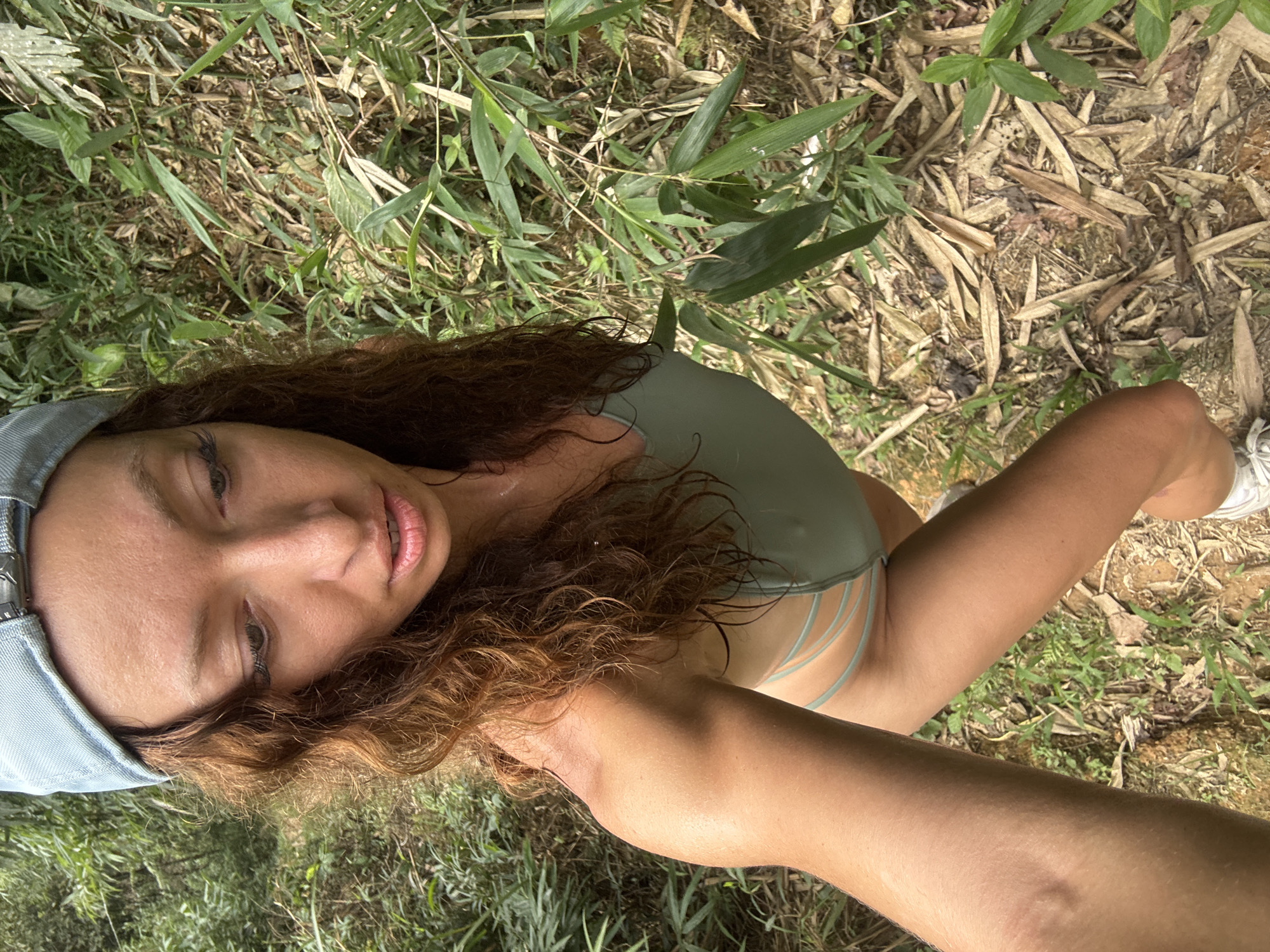 The image presents a woman in a natural setting. The surrounding environment includes grass, leaves, and what appear to be bamboo leaves scattered on the ground. She is wearing a light blue cap and a light green swimsuit. Her hair is curly and reddish-brown. The angle suggests the photo was taken from a low perspective. The exact location within Suối Giàng cannot be determined from the visual information provided.