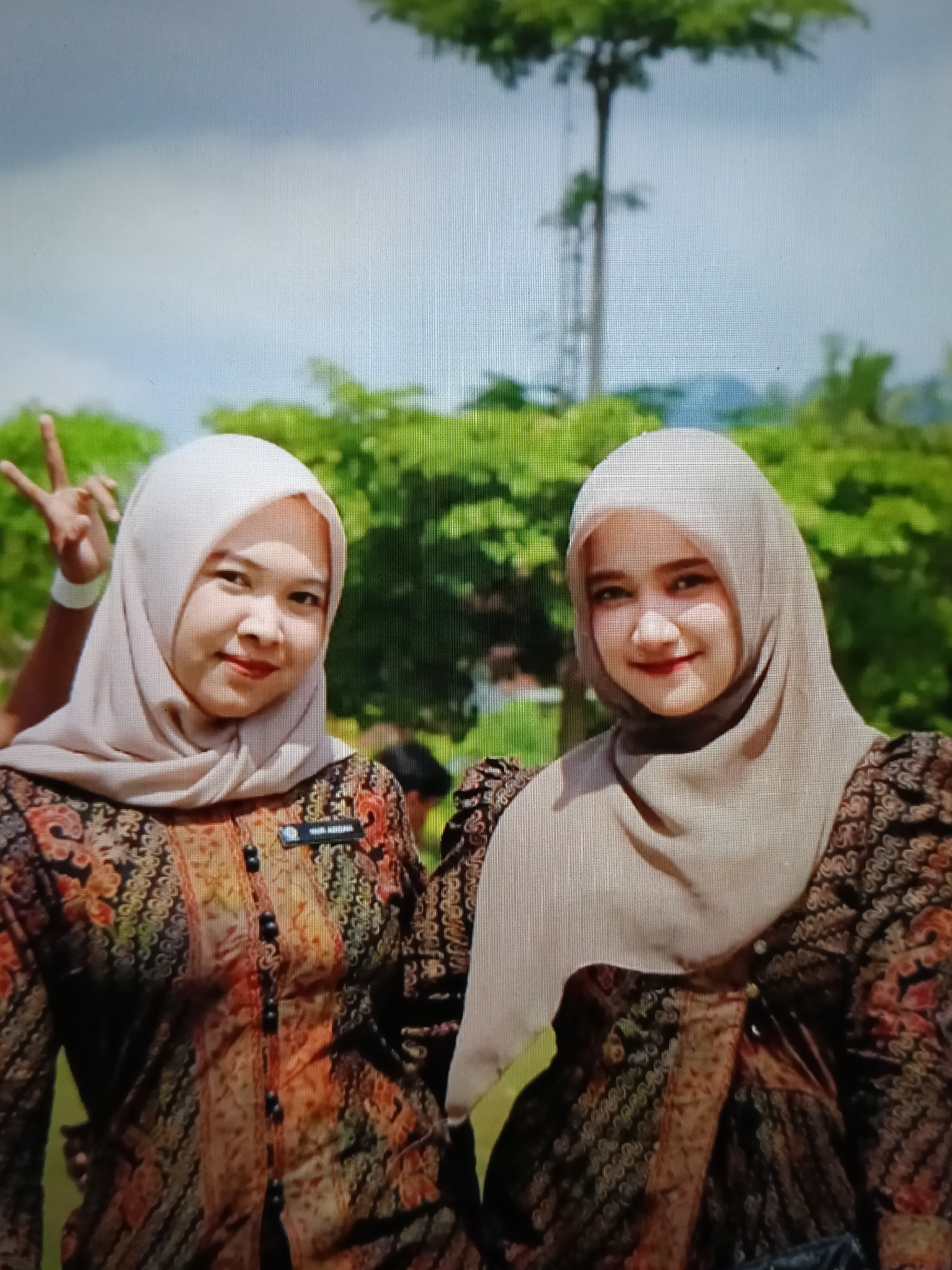 The image shows two young women posing for a photo outdoors. They are both wearing headscarves and batik patterned tops. The woman on the left is wearing a light brown headscarf and a multi-colored batik shirt with a name tag that reads "DWI AMALIA". She is making a "peace" sign with her fingers. The woman on the right is wearing a similar light brown headscarf and a dark batik shirt. Both women are smiling. In the background, there are green trees and a cloudy sky. The overall setting appears to be a park or outdoor recreational area.