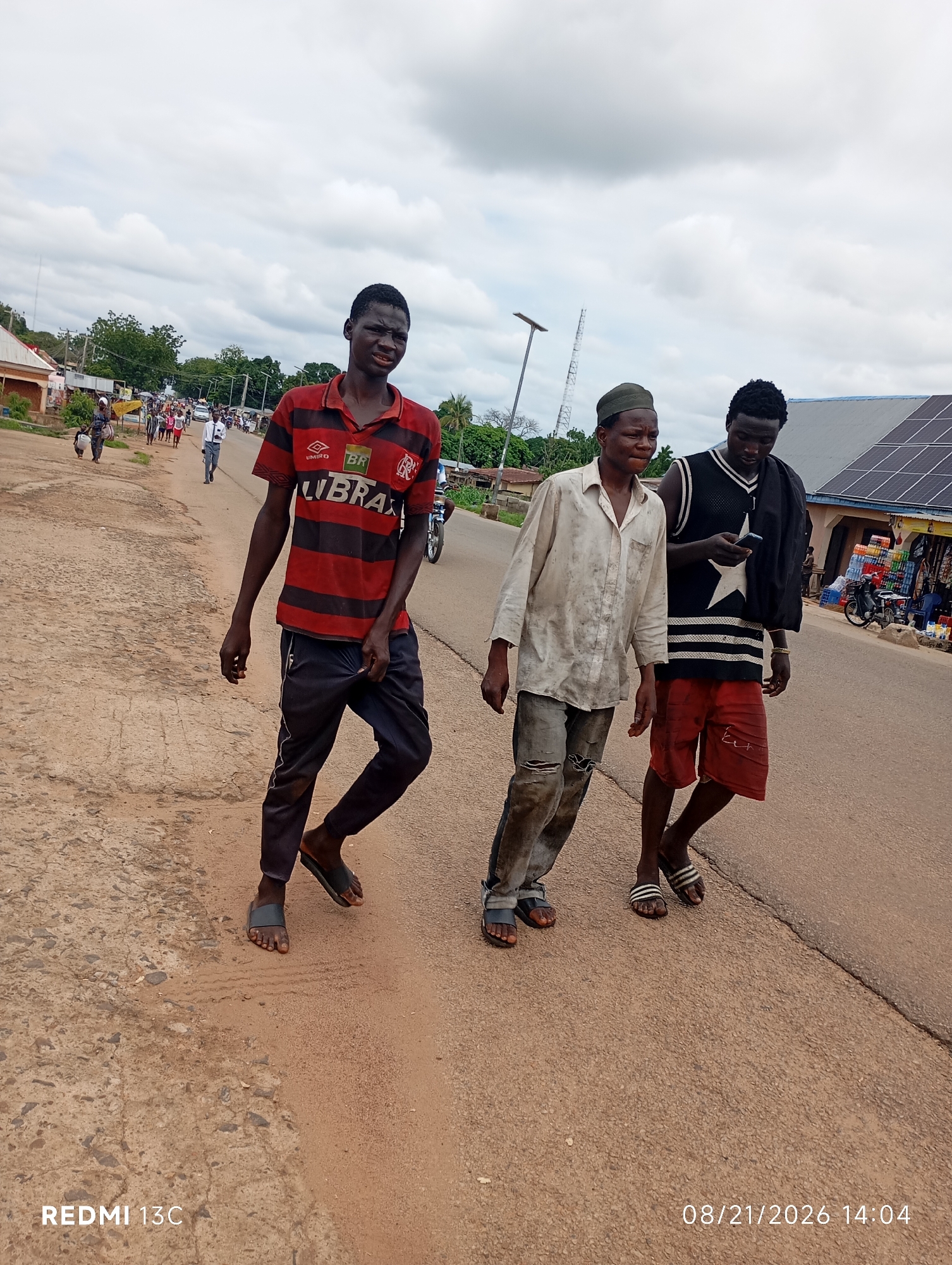 The image shows three young men walking on a dusty road in the city of Shendam, Nigeria. The man in the foreground is wearing a red and black striped t-shirt and dark pants. The man in the middle is wearing a light-colored, dirty shirt and ripped pants, with a green cap on his head. The man on the right is wearing a black tank top with a white star and red shorts, and is holding a smartphone. The background shows buildings, solar panels, trees, and other people on the side of the road. The sky is cloudy. The date stamp on the image indicates it was taken on August 21, 2026, at 2:04 PM.