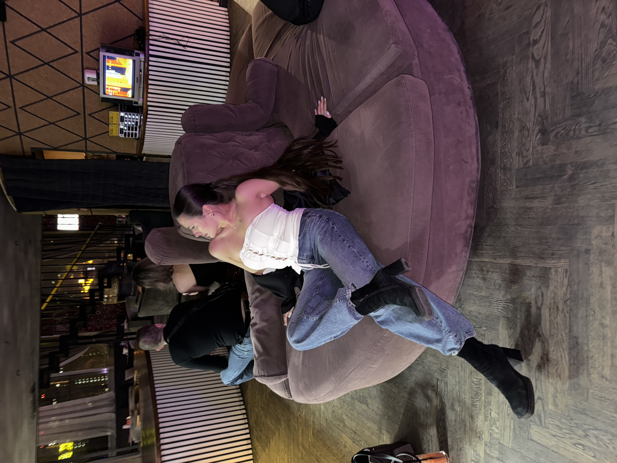 The photograph depicts a social gathering in what appears to be a bar or lounge setting. A woman with long dark hair is reclining on a large, circular, plush sofa, dressed in a white top, light blue jeans, and black heeled boots. Another person, wearing a black shirt, is partially visible next to her on the sofa. A man wearing a dark jacket is seated on the sofa as well.
In the background, there is a television set playing content. The interior design features dark wood flooring and a black and white striped wall. Outside the window, it is apparent that this is a city setting, likely Moscow, Russia, given the urban skyline visible through the glass windows.