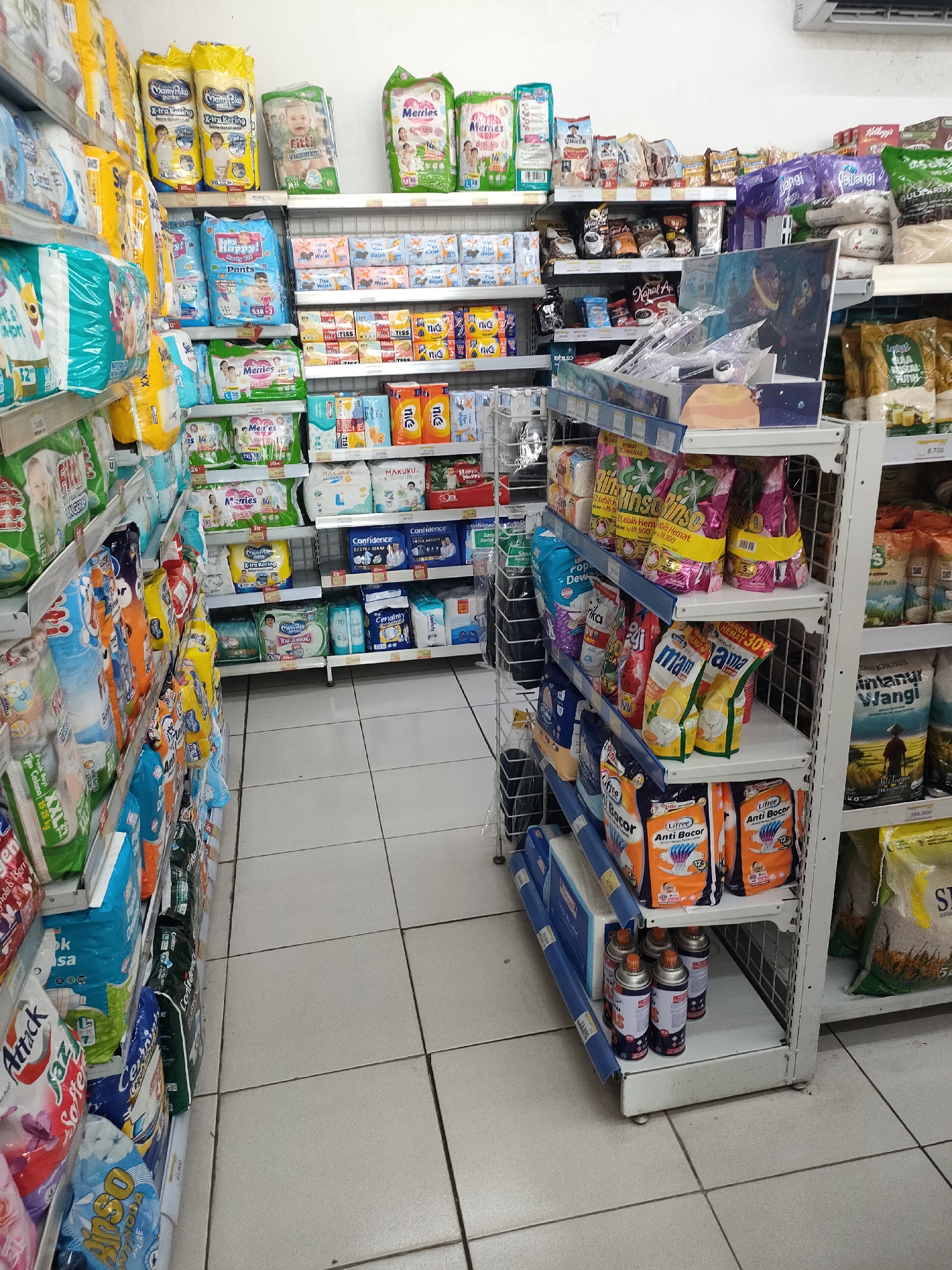 This is a forensic, matter-of-fact description of an indoor scene, likely a convenience store or supermarket aisle in Curahdringu, Indonesia. The image captures a view of shelves stocked with various consumer products, primarily packaged goods such as diapers, baby wipes, laundry detergent, and possibly food items.
The shelves are constructed of white metal, arranged in vertical tiers. On the left side of the frame, several shelves are densely packed with large, colorful plastic packages of diapers. Brands and designs vary, featuring images of babies and text in Indonesian. Some packages are turquoise and white, others yellow and white, and some are red and white. Further down the aisle, on the right side, more shelves display a different assortment of products. These include smaller packets of what appear to be facial tissues or wet wipes, and larger bags of what could be rice or grains, one with green packaging and an image of rice stalks. There are also canisters, possibly air fresheners or cleaning sprays, on a lower shelf towards the bottom right.
The aisle itself is narrow, with white tiled flooring that shows subtle grout lines. The lighting appears to be artificial, casting a bright, even illumination throughout the space. There are no people visible in the frame. The overall impression is one of a well-stocked retail environment, organized for product display and customer access. The presence of specific product packaging with Indonesian text suggests a local context within Curahdringu, Indonesia.