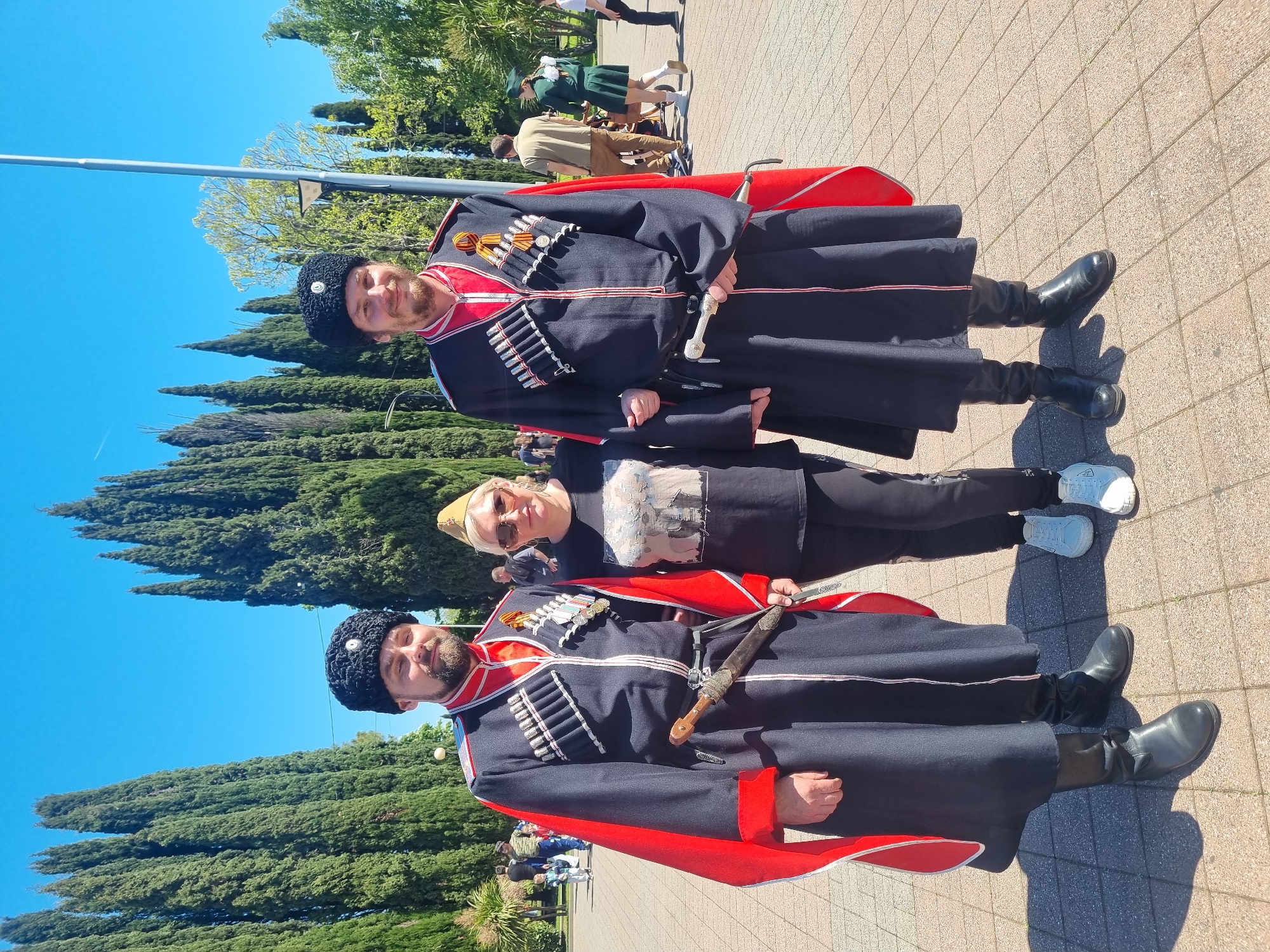 Two men in traditional Caucasian clothing and black boots pose outdoors with a woman on a paved surface. The men are wearing black wool hats, black coats with red trim, decorative silver accents, and daggers. The woman wears a black t-shirt, black pants, and white sneakers. The background includes tall green trees and a clear blue sky. The location is likely in Sochi, Russia.