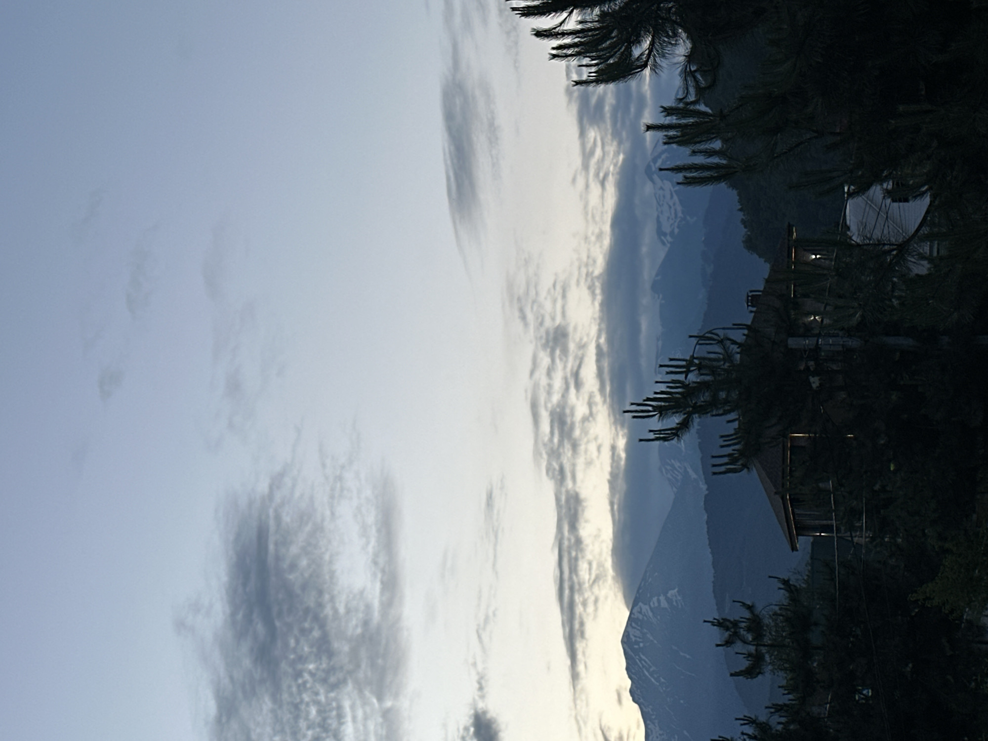 A twilight landscape photograph taken from a high vantage point in Krasnaya Polyana, Russia, depicts a serene mountain vista. In the foreground, dark silhouettes of pine trees frame the scene, with their needles sharp against the pale sky. A small, rustic wooden house with a dark roof is visible amidst the trees, its windows softly illuminated. Beyond the trees and house, layers of forested hills recede into the distance, leading to imposing, snow-capped mountains under a pale blue sky dotted with wispy clouds. The overall impression is one of a quiet, natural setting at the edge of dusk.