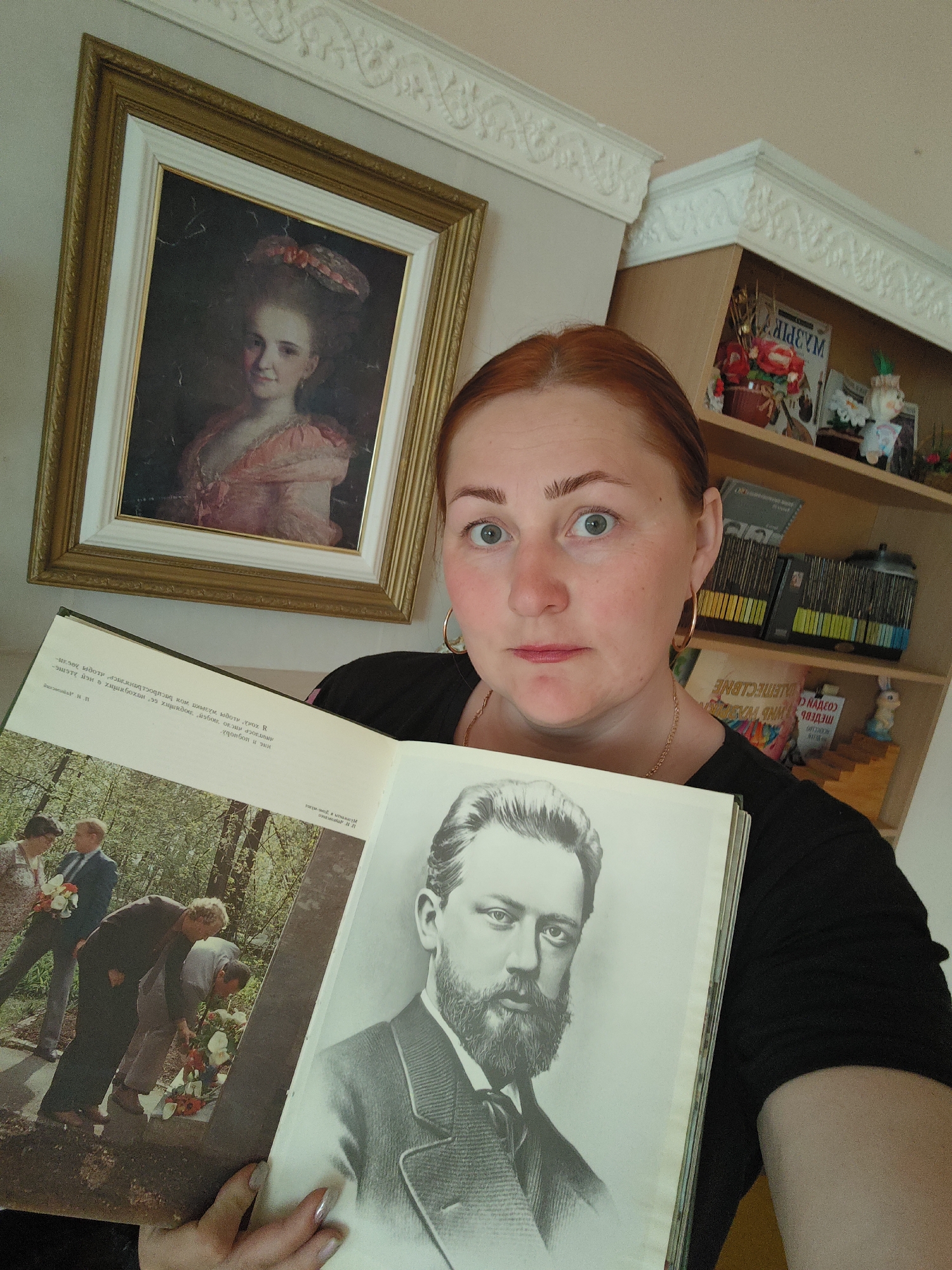A fair-skinned woman with red hair and gold hoop earrings is holding a book open in front of her. The book has a black and white portrait of a man with a beard on the right page, and a color photo of people placing flowers on a grave on the left. Behind the woman is a framed portrait of a woman in a pink dress. To the right of the woman is a bookcase with various items on the shelves. The wall and ceiling have decorative molding. The OCR in the image suggests the location is possibly connected to Markovskii, Russia.