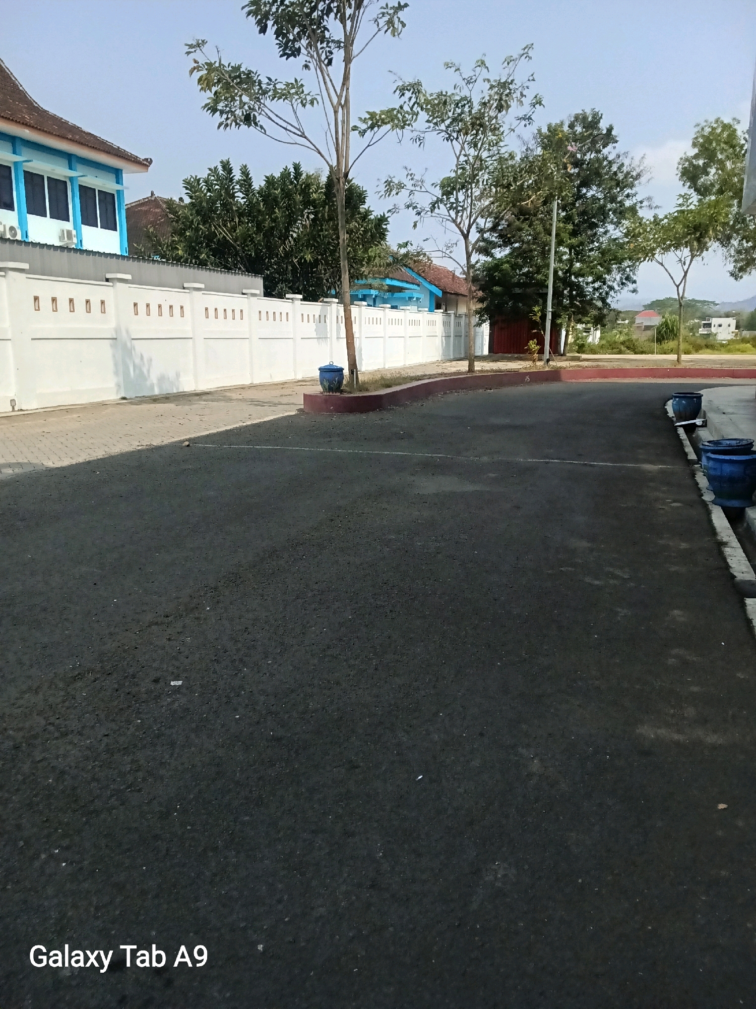 The image depicts an outdoor scene, likely a parking area or courtyard, under a clear blue sky. In the background, a light blue building with white trim and dark windows is visible, partially obscured by trees. A white decorative wall with square cutouts runs horizontally across the mid-ground. Several trees of varying sizes are scattered throughout the scene, with their branches reaching towards the sky. The ground in the foreground is paved with dark asphalt. Two blue ceramic planters are situated near the wall, one on the left and another on the right side of the image. Further in the distance, more buildings and vegetation can be seen, suggesting a semi-urban or suburban environment. The lower left corner of the image displays the text "Galaxy Tab A9". The image was taken in Trenggalek, Indonesia.