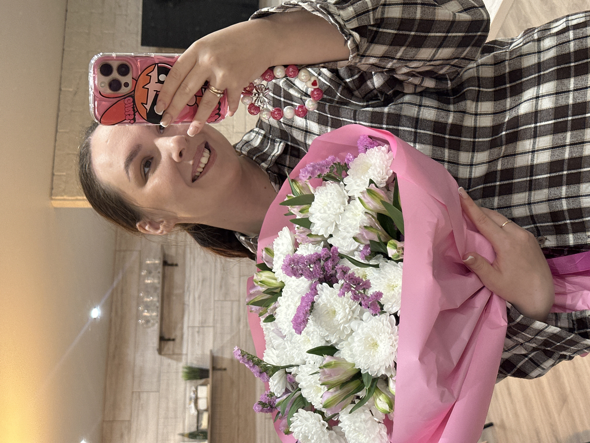 A woman is depicted taking a selfie indoors, smiling broadly at the camera. She is wearing a brown and white plaid long-sleeved shirt. Her dark hair appears to be pulled back.
In her left hand, she holds a smartphone with a clear pink case that features a cartoon character, possibly Bubbles from The Powerpuff Girls, identifiable by its large orange hair. The phone's triple camera lens setup is visible. She wears two rings on her left ring finger, and her fingernails have a French manicure style.
Her right arm supports a large, vibrant bouquet of flowers wrapped in bright pink paper. The bouquet predominantly features numerous white chrysanthemum-like flowers, interspersed with smaller clusters of purple statice flowers and some light green/white lilies or alstroemeria that are not fully bloomed. On her right wrist, she wears a bracelet composed of alternating red/pink and white beads, accented with a silver-toned charm.
The background shows an indoor environment. A plain light-colored wall is visible on the left, while the right side features a wall with vertical light wood-like paneling or tiles. In the distance, shelves adorned with small plants and other decorative items can be seen. Bright reflections from light sources are present on the left wall. The woman's joyful expression suggests happiness. There is no visible text in the image. The scene is set in Chaikovsky, Russia.
