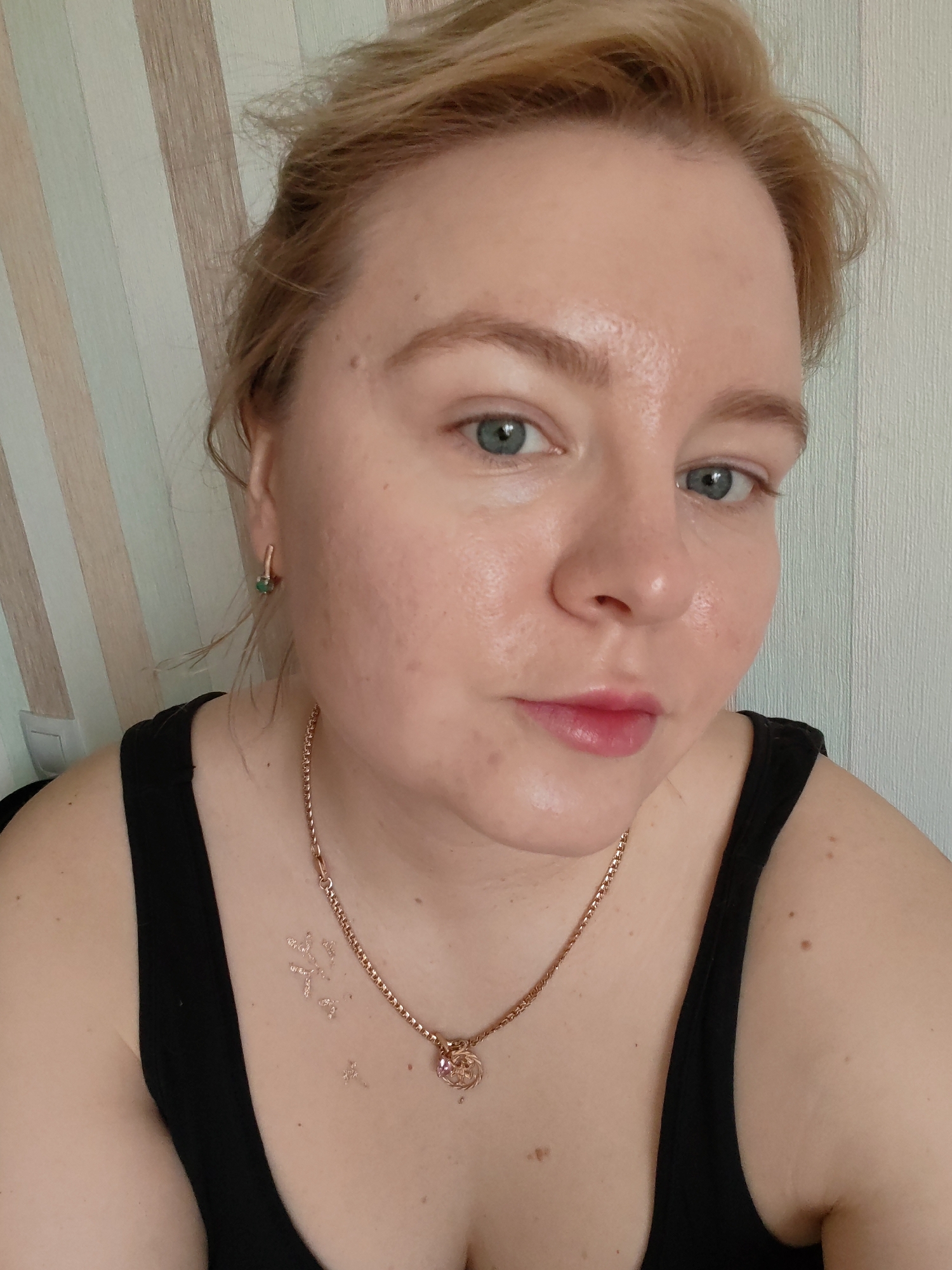 A light-skinned woman with blonde hair is seen in a close-up, self-portrait style photograph. She is wearing a black tank top and a gold necklace with a pendant. She has light eyes and lightly colored eyebrows. A small green earring is visible on her left ear. There are small, seemingly temporary, tattoos on her chest. The background features a vertically striped wall in light shades of brown, white, and light green. There is no information present in the image indicating that it was taken in Izhevsk, Russia.