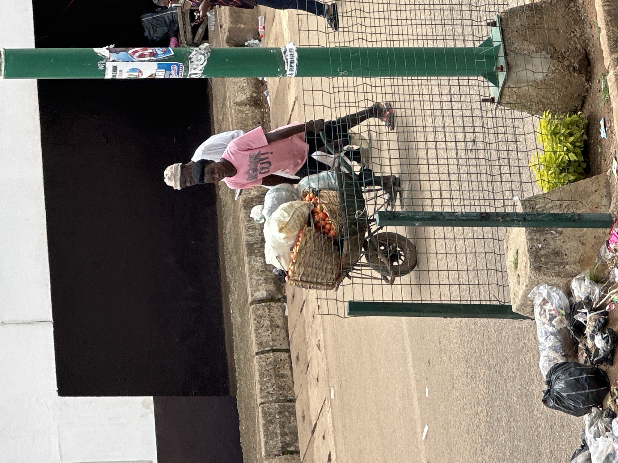 The image depicts a street scene in Bukuru, Nigeria. In the foreground, a wire mesh fence encloses a concrete base supporting a green metal pole. To the left of the fence, a pile of refuse, including black plastic bags and other debris, is visible on the ground.
Behind the fence, a wheelbarrow filled with produce, primarily tomatoes, and some white cloth bags is being pushed by a young person wearing a pink t-shirt and dark pants. Another person, an older individual wearing a white shirt and a light-colored cap, walks behind the wheelbarrow, appearing to be accompanying it. They are on a paved surface with a concrete curb bordering a darker paved area.
In the background, a building with a white upper section and a dark lower section is visible. A green utility pole stands to the right of the frame, adorned with several posters. A person in a red and black checkered shirt and dark pants is seen walking on the darker paved area in the background, holding a mobile phone. The overall environment suggests a bustling urban street, likely a market or a thoroughfare where goods are transported.