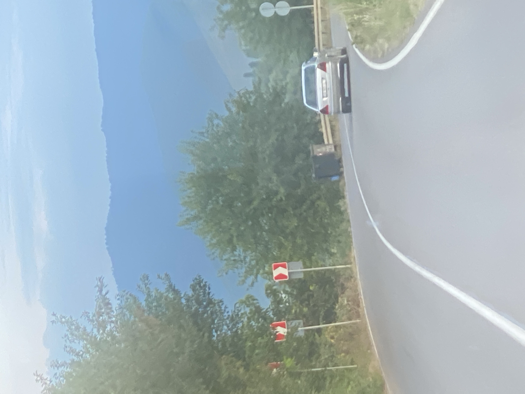 The image captures a serene, sunlit day in Zemo Sarali, Georgia. A silver sedan navigates a gentle curve on a paved road, its rear clearly visible. The road is lined with lush green trees, and in the distance, rolling hills are softened by a hazy blue sky. Road signs are strategically placed, indicating the road's contour, and two trash bins stand near the roadside. The overall atmosphere is one of tranquil, rural charm.