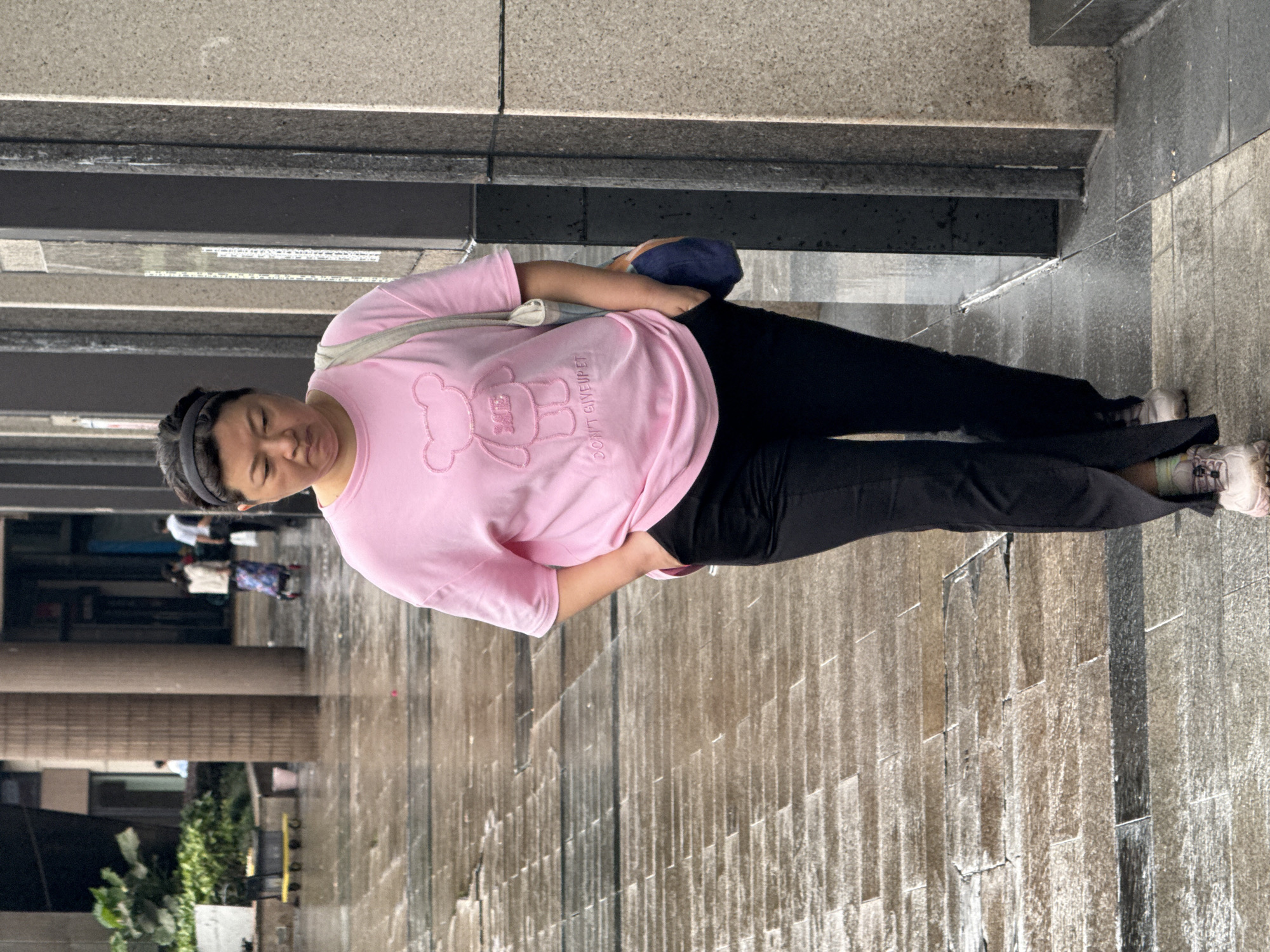 A woman stands on a gray-tiled walkway in the Nan'an District of China. She wears a pink long-sleeved shirt with a bear graphic and the phrase "DON'T GIVE UP PET," black pants, and light-colored sneakers. A dark headband is visible in her hair, and she carries a light-colored bag over her shoulder and a blue object, possibly a hat, in her hand. The walkway features vertical, textured tile patterns. In the background, a building with columns and greenery is partially visible. Other people can be seen walking in the distance. A dark, overhanging structure appears above the woman.