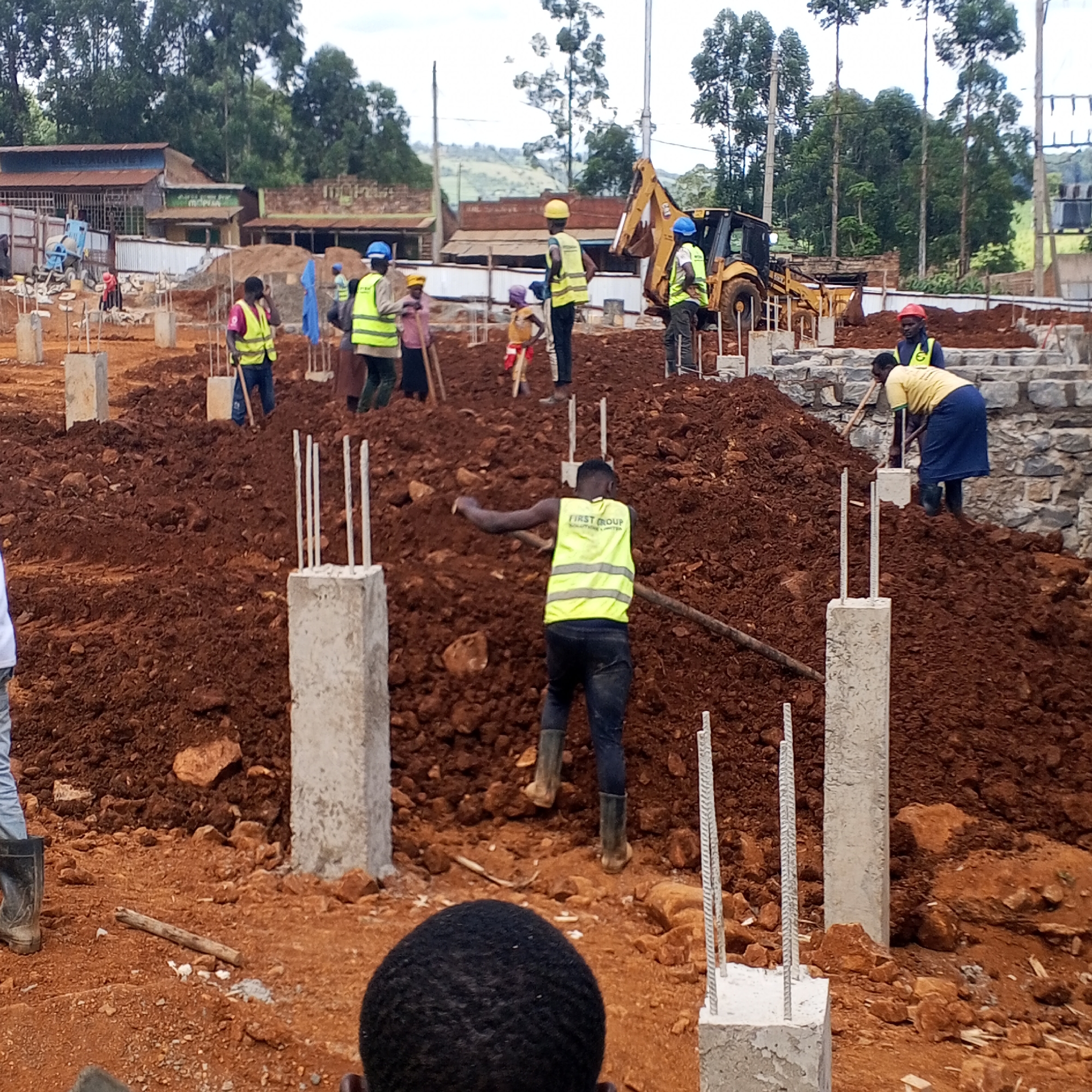 An outdoor construction site in Borabu Chitago ward, Kenya, is depicted during daylight hours under an overcast sky. The scene is dominated by reddish-brown earth, uneven mounds of soil, and newly set concrete foundation pillars with exposed rebar.
Numerous individuals, primarily adults, are engaged in construction work. Many are wearing high-visibility vests (yellow or green) and some have hard hats (blue, yellow, red), indicating safety consciousness. In the foreground, a worker seen from behind wears a yellow vest emblazoned with "FIRST GROUP SOLUTIONS LIMITED" and appears to be working with a tool, possibly a shovel. Another worker, a woman in a yellow shirt and blue skirt, is actively shoveling soil next to a partially constructed stone retaining wall in the midground. Other workers are distributed throughout the site, some holding shovels or hoes, suggesting manual earthmoving and leveling. Two children are also visible among the workers, one in a yellow shirt holding a stick.
A yellow backhoe loader is positioned in the mid-right background, implying mechanical assistance for larger earthworks. A blue cement mixer is visible in the left background. The distant background features simple, single-story buildings with corrugated iron roofs, surrounded by lush green trees and hilly terrain, characteristic of a rural or semi-urban environment in Kenya. The soft lighting suggests cloudy weather. The image captures the active groundwork phase of a building project, emphasizing human labor alongside some machinery.