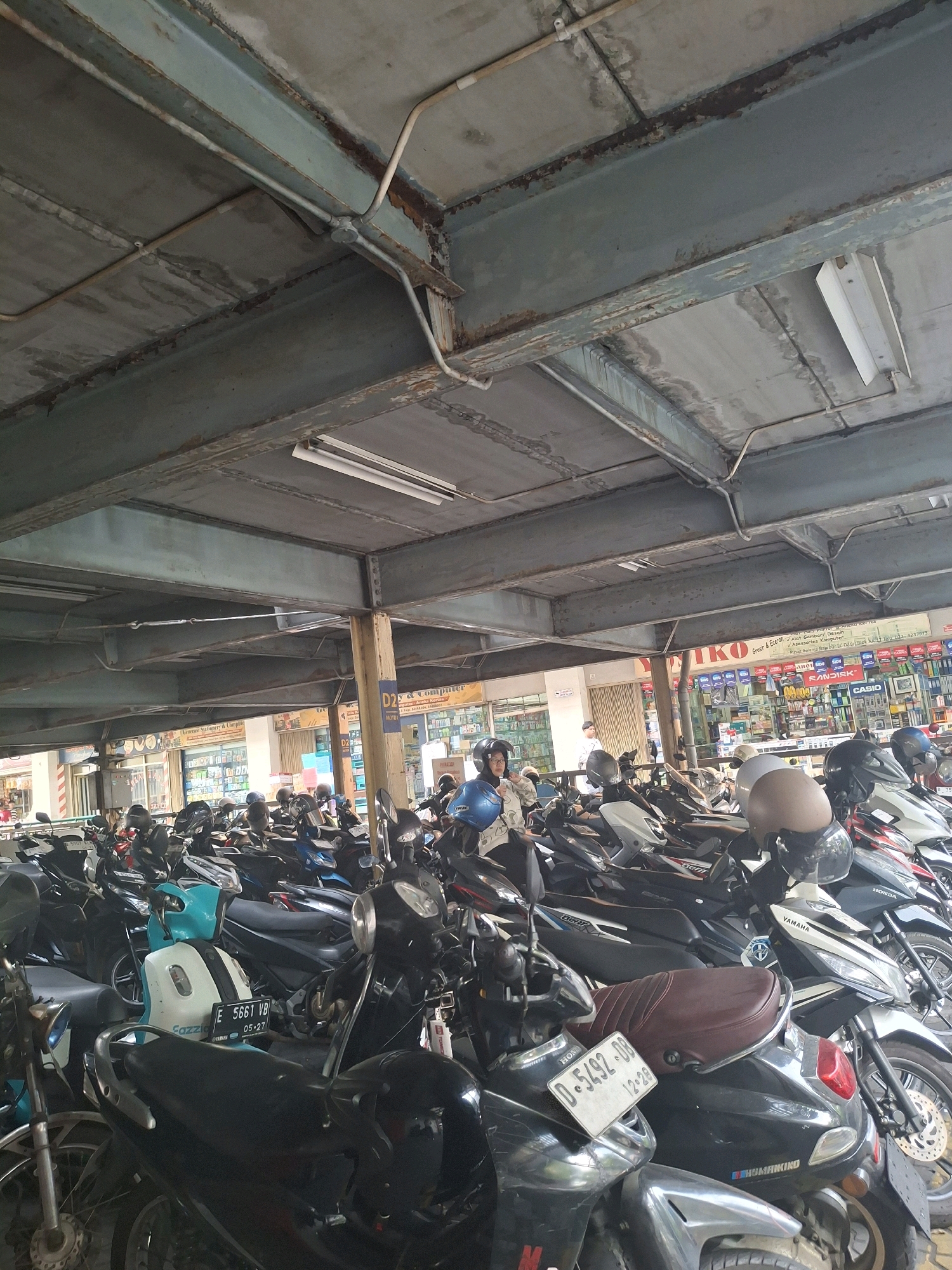 The image depicts a crowded motorcycle parking lot under a covered structure, likely in Bandung City, Indonesia. The structure's ceiling is made of concrete panels supported by weathered steel beams, with exposed electrical conduits running overhead. Fluorescent lights are mounted on the ceiling. The parking lot is filled with numerous motorcycles of various models and colors, parked closely together. Some motorcycles have helmets placed on them or nearby. In the background, there are storefronts with signs, suggesting the parking lot is adjacent to shops or a commercial area. One prominent sign reads "D2Y & Computer." Several people are visible, some wearing helmets and others walking. One woman in the foreground is wearing a blue helmet and a patterned shirt, appearing to be checking something on her motorcycle. Another person in the background is wearing a white shirt and dark pants. The overall impression is a busy urban environment with a focus on motorcycle transportation.