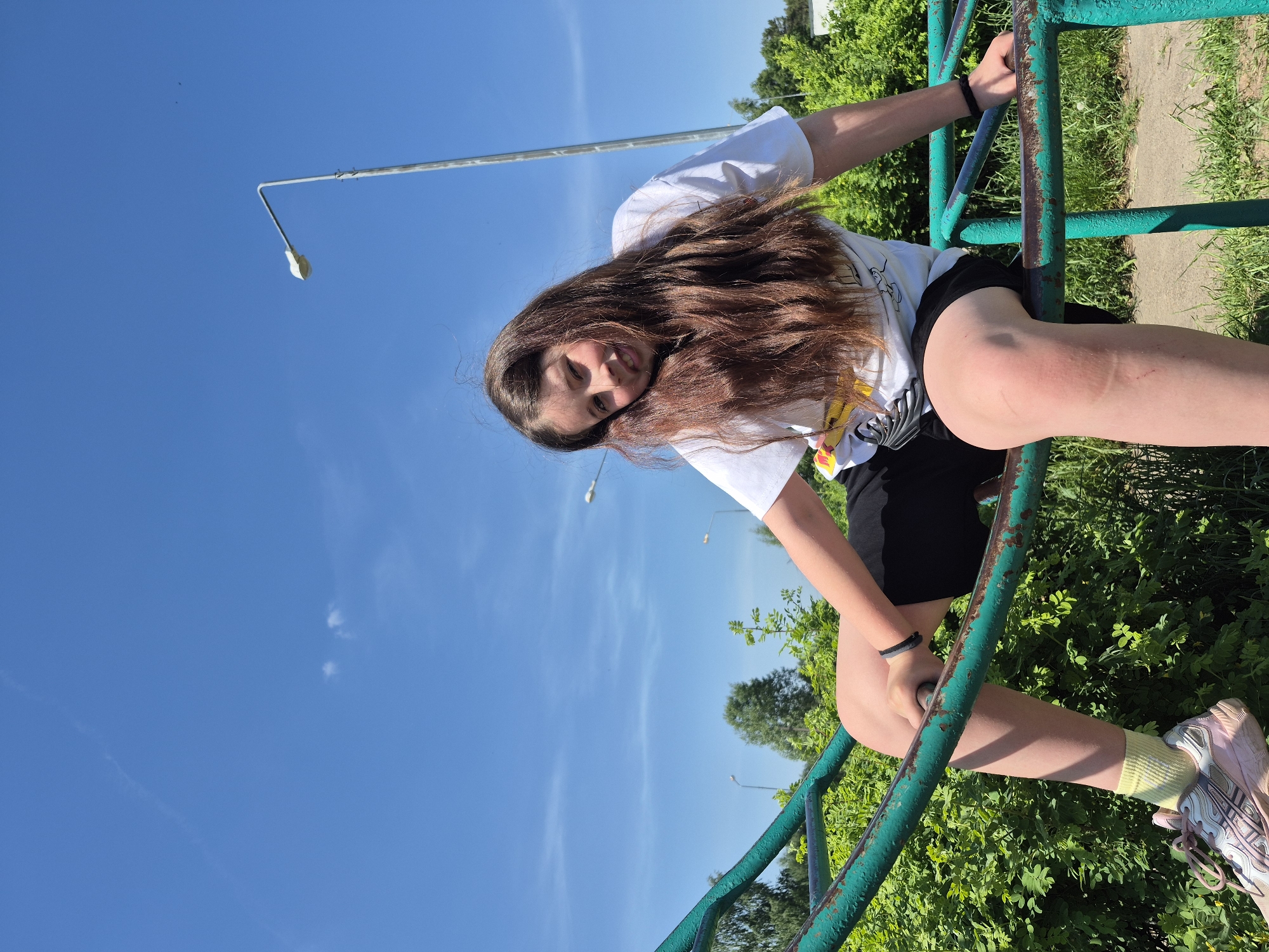 A girl with long brown hair, wearing a white t-shirt and black shorts, is hanging on a green and blue metal structure. She is smiling towards the camera. Her legs are extended and one foot is wearing a yellow sock and a pink and grey athletic shoe. The background features a clear blue sky with scattered clouds. A tall streetlight is visible in the distance, along with green trees and grass. The scene appears to be in a park or recreational area.