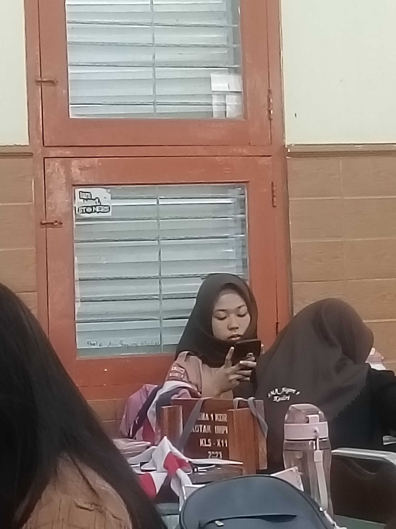 The image depicts a group of young people, likely students, in what appears to be a classroom or study area within Kediri City, Indonesia. Two individuals are prominently visible in the foreground. On the left, a young woman wearing a hijab is focused on her mobile phone, holding it in her hands. Her facial features are visible, and she is wearing a light-colored hijab. To her right, another person, also wearing a hijab, is partially obscured. Their head is bowed, and they are facing away from the camera, with their right shoulder and the side of their head visible.
In the background, a wooden cabinet with glass doors is visible. The glass panes have blinds behind them, suggesting a window or partition. A sticker with text and a logo is affixed to the lower section of the cabinet door. There are stacks of papers or folders on a table or desk in front of the students, with some items of clothing, possibly scarves or fabric, draped over them. A water bottle with a pink lid and a white strap is also present on the table. The overall environment appears to be indoors, with neutral-colored walls and a wooden floor or tiled surface. The lighting suggests it might be daytime, but the image quality is somewhat low, making it difficult to ascertain finer details about the surroundings. The scene conveys a sense of quiet study or a break between activities.