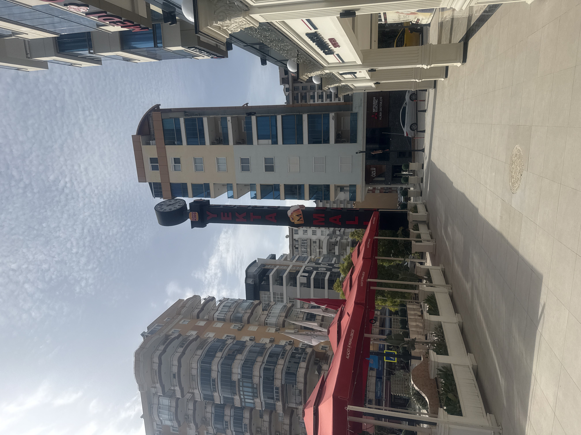 The image captures an outdoor scene at Yekta Mall in an unidentified city. The perspective is rotated, making the buildings appear vertical. Buildings are situated on either side of a pedestrian walkway. One building is a high-rise apartment complex with multiple floors and a curved façade. Another building is light-colored, with a rectangular shape and a clock near the top. A tall Yekta Mall sign stands in the foreground. Red awnings are visible. On the right side of the walkway, there are planters and architectural columns. A white car is parked near a building with a Mitsubishi Electric sign. The sky is lightly cloudy.