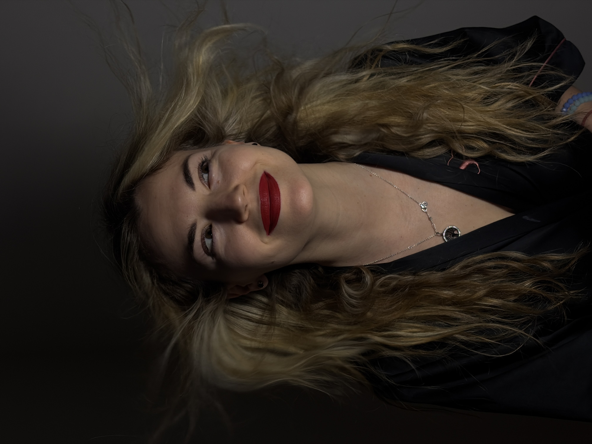 A low-angle, medium shot depicts a woman lying on a dark surface. The woman has light blonde hair with darker roots, fair skin, and wears red lipstick. She is wearing a black garment and a silver necklace with a circular pendant. The background is a dark, indistinct gray or black. The setting appears to be indoors. The location, Budva, Montenegro, cannot be verified based on the image content.