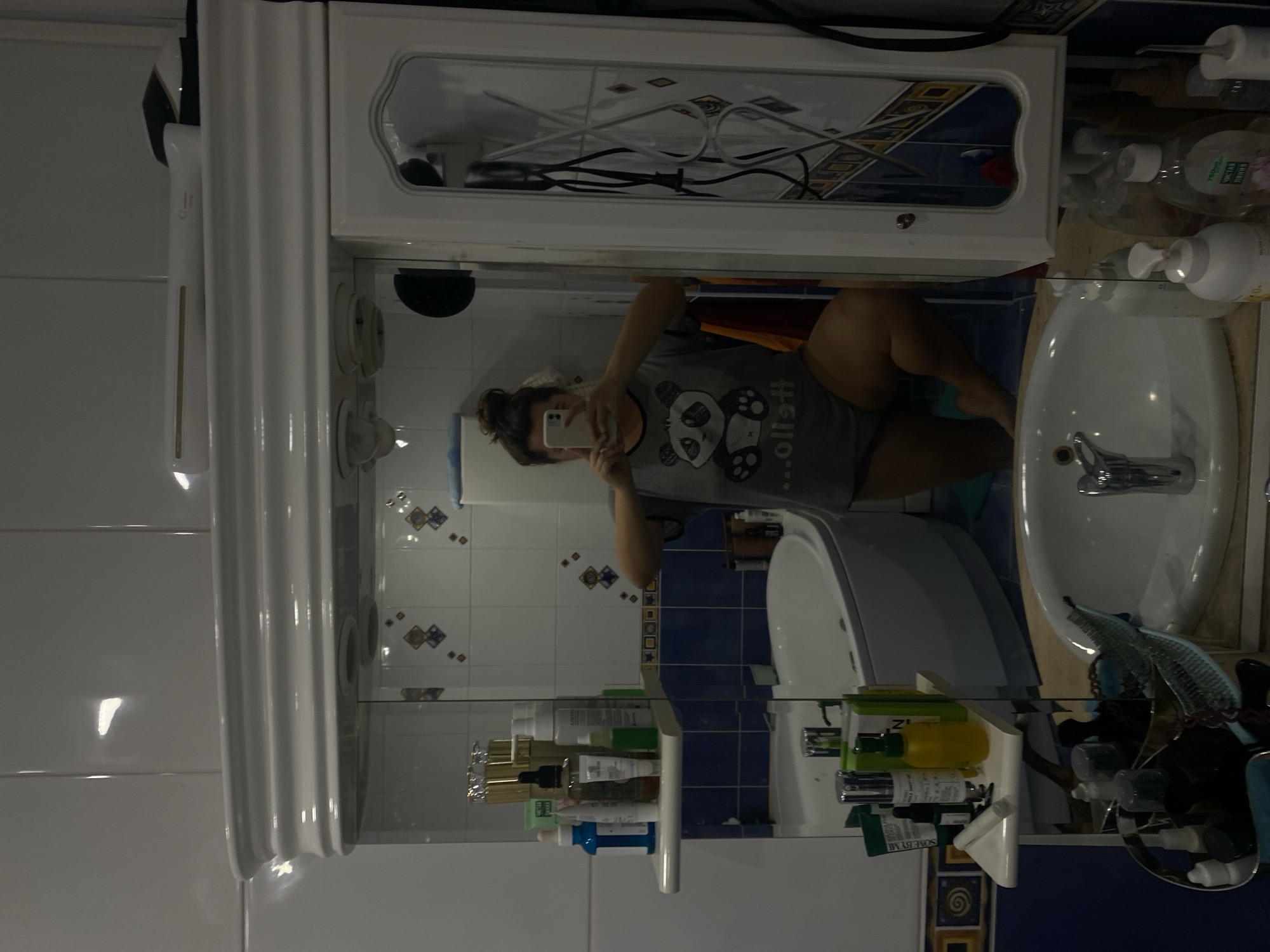 This image shows a woman taking a selfie in a bathroom, reflected in a mirror. She is wearing a grey t-shirt with a panda design and dark shorts, and is kneeling on the floor. The bathroom appears to be in a residential setting. The mirror reflects various items, including toiletries on shelves, a bathtub, a sink, and a medicine cabinet. The tiles on the wall are white and blue, with some decorative elements. The overall impression is of a personal, everyday moment captured in a private space. No information about the city of Perm, Russia is present in the image.