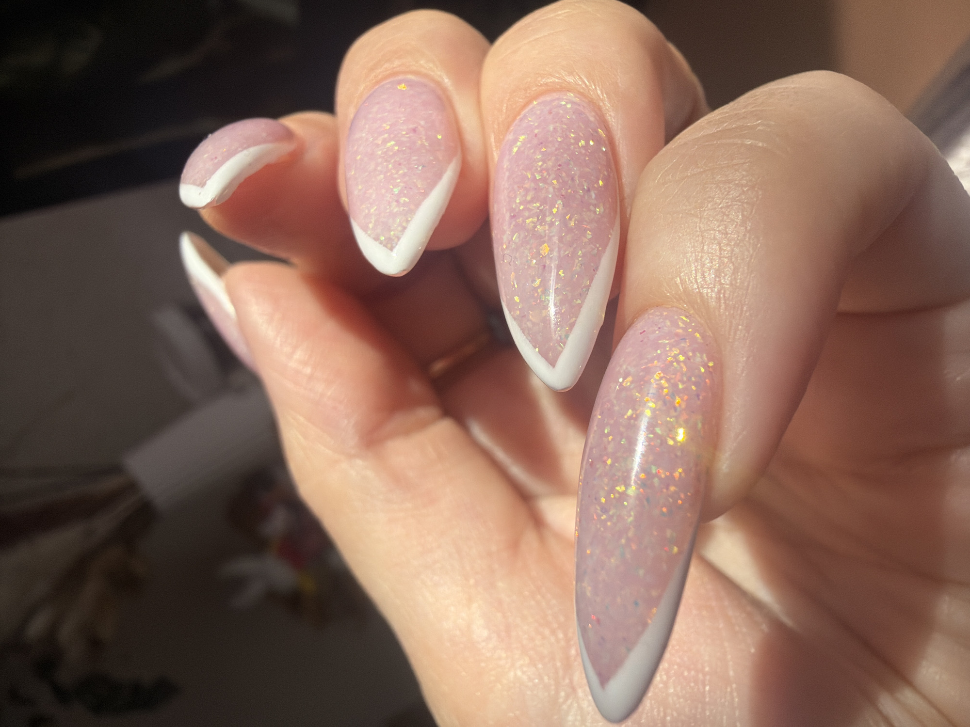 A close-up photograph displays a person's hand featuring several fingernails with manicured, almond-shaped tips. The nails exhibit a pale pink base color interspersed with glitter or shimmering particles. A crisp white line accentuates the shape of the nail tips in a geometric V-shape. The skin appears smooth and healthy. The background is out of focus, displaying indistinct objects and shadows. The location is not deducible from the image.