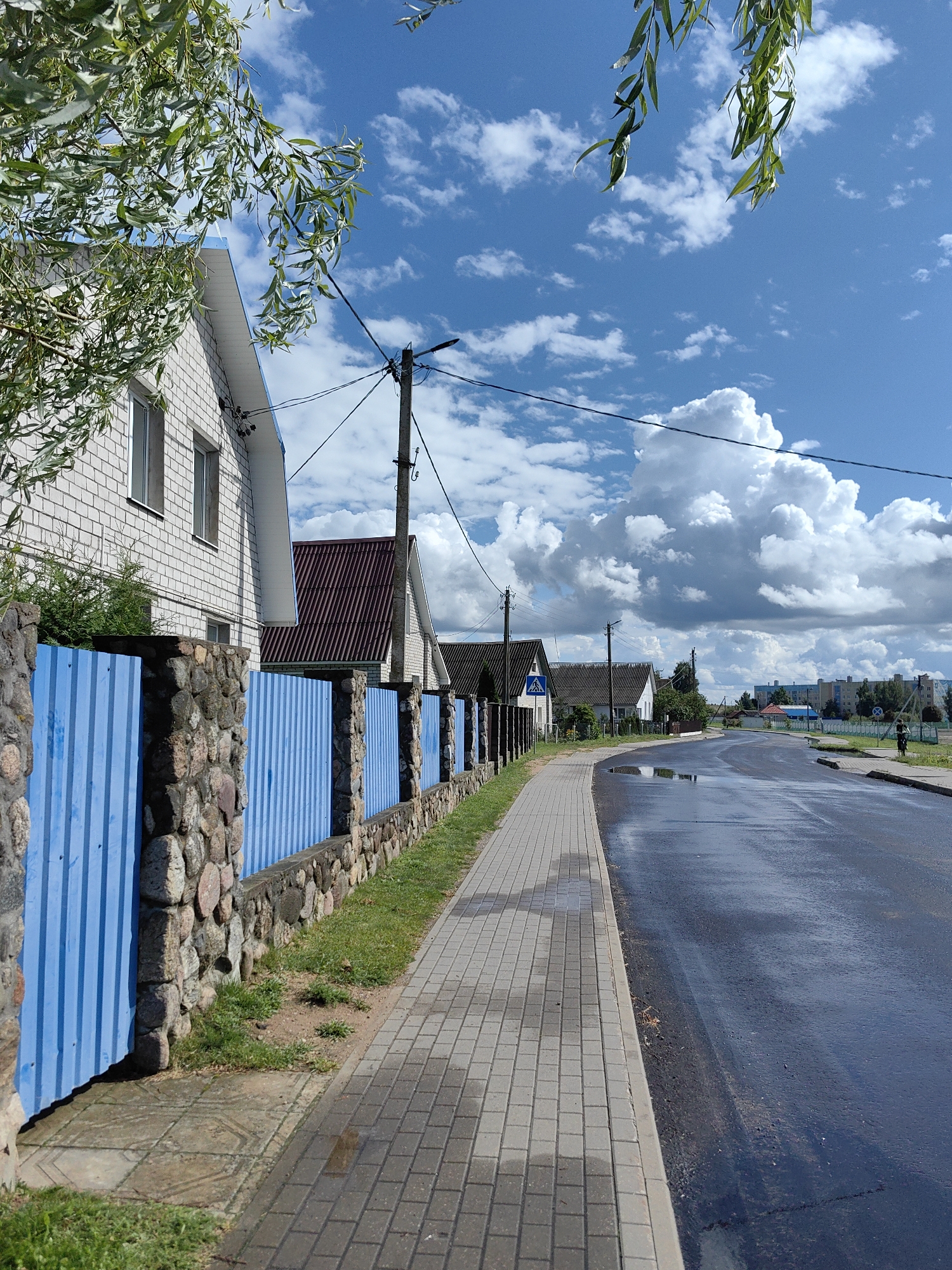 The image depicts a street scene in Myadzyel, Belarus, on a partly cloudy day. On the left, a light-colored brick house with a dark roof is partially visible, bordered by a fence composed of stone pillars and blue corrugated metal panels. The fence runs along a paved sidewalk made of gray rectangular bricks. The sidewalk is wet in places, reflecting the sky.
The road to the right of the sidewalk is dark and also wet, with visible puddles reflecting the sky and clouds. Several utility poles with electrical wires are present along the street. Further down the road, a few more houses with similar dark roofs are visible, spaced apart. In the distance, there are apartment buildings and trees, indicating a more urban or suburban area. A person is walking in the distance on the right side of the road. The sky is a vibrant blue with scattered, fluffy white clouds, suggesting fair weather. The leaves of a tree hang down from the top left corner of the image, framing the scene.
