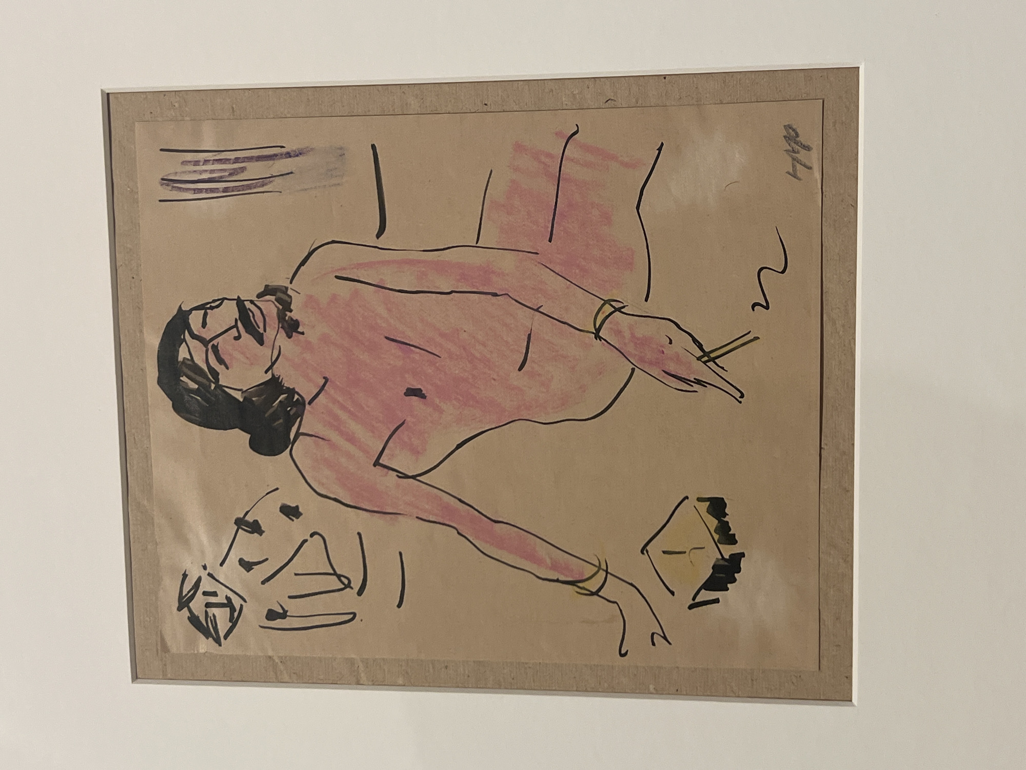 A framed sketch depicts a reclining nude figure, rendered in black outline with pink shading, on a tan background. The figure has dark hair and appears to be wearing glasses. The figure is holding what may be a cigarette. There are abstract shapes at the bottom left and right. There is some writing in the top right of the sketch. The location is not identifiable as Tel-Aviv, Israel.