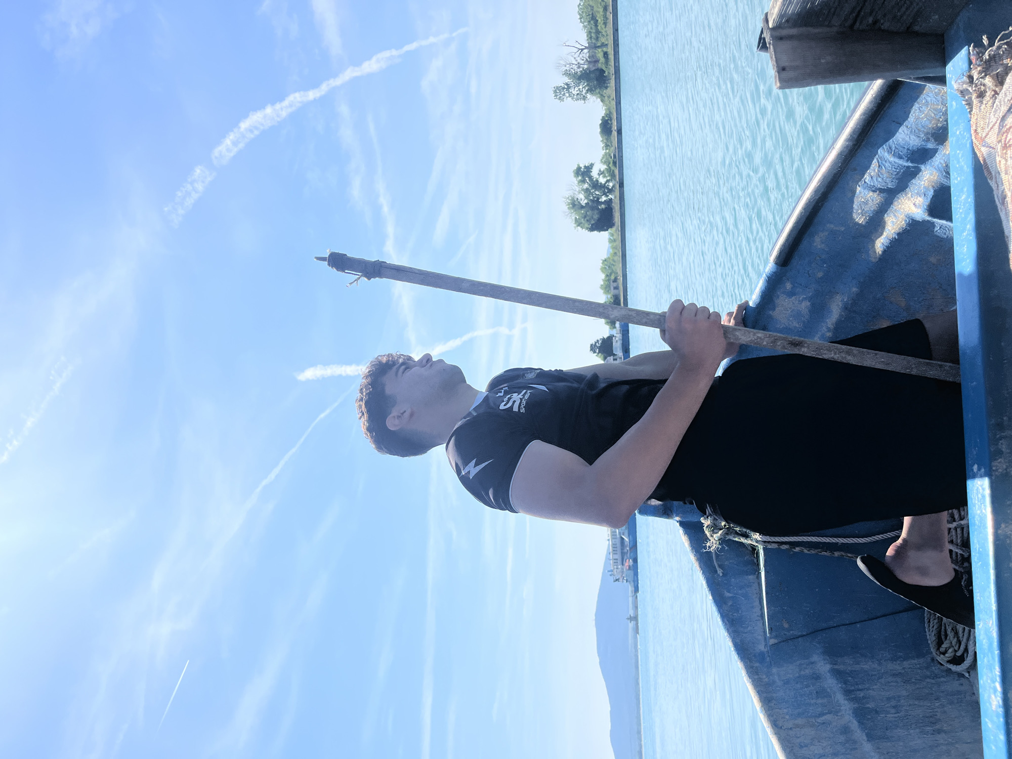 The location is unknown. A young man stands in a weathered blue wooden boat on calm, light blue water under a clear, bright blue sky. He is seen from the chest up, with a partial profile facing towards the viewer's right, looking towards the horizon. He has short, curly dark hair and is wearing a black t-shirt with white lightning bolt designs on the sleeves and a "SPORTBEAR" logo on the chest. He wears black shorts, and his right foot, partially visible, is bare and resting in a black flip-flop or sandal. The man holds a long, vertical wooden pole with both hands; the top of the pole has twine or fabric wrapped around it, resembling a rudimentary torch. The interior of the boat is a faded, peeling blue, with coiled ropes visible near the man's feet and a patterned textile on the bottom right corner. In the distant left background, a large mountain or hill is visible under the horizon, alongside a light-colored ship or ferry and a small structure near the shore. The right background features a flat coastline with scattered green trees. The bright blue sky is marked by multiple white contrails from aircraft. The scene is brightly lit by natural sunlight, creating reflections on the water.