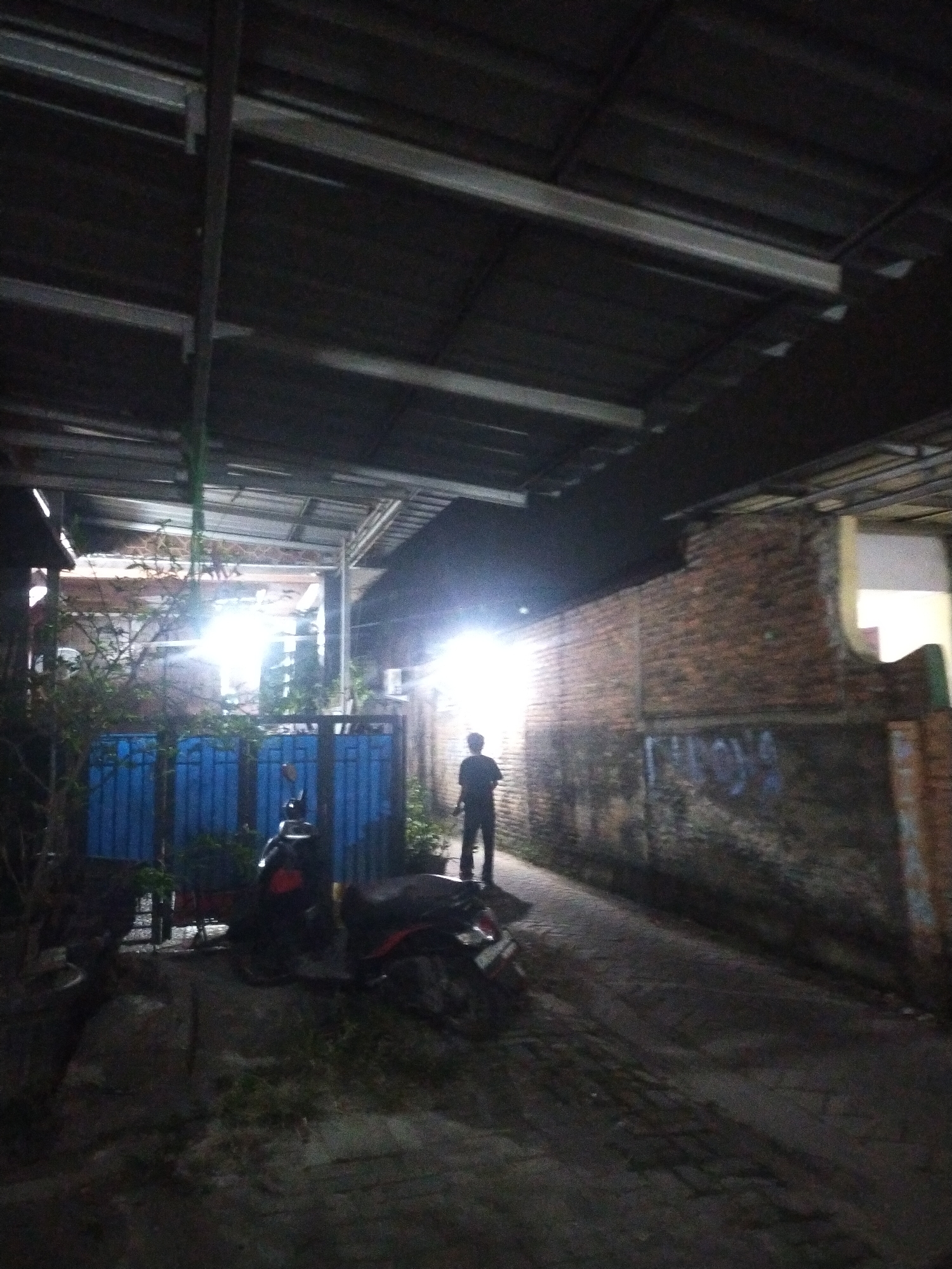 The image depicts a dimly lit alleyway in Kampung Besar, Indonesia. A solitary figure stands in the middle of the alley, facing away from the camera. The figure is silhouetted against a bright light source originating from a building in the background. To the left of the alley, there are blue gates and some greenery. Two motorcycles are parked in the alley. The walls of the alley are constructed of brick. The scene appears to be at night.