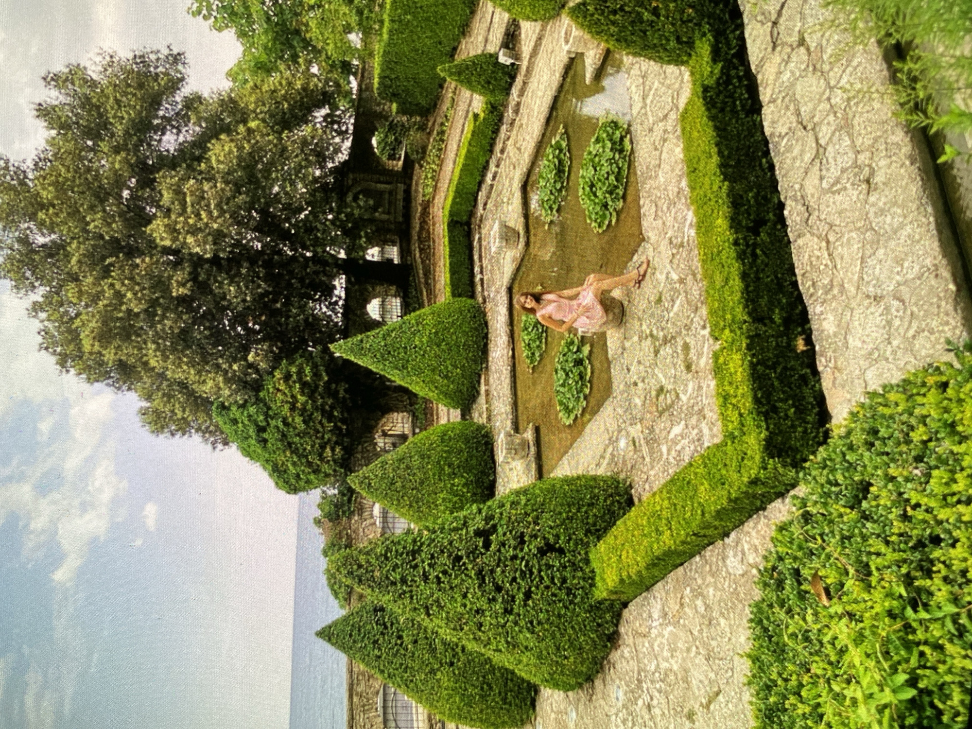 The image, oriented on its side (rotated 90 degrees counter-clockwise), captures a serene and formal garden scene under a bright, partly cloudy sky. With a provided location context of Bankya, Bulgaria, the setting features meticulously maintained topiary and stone architecture.
In the foreground and midground, a formal garden unfolds, characterized by precisely shaped green hedges, including cone-shaped topiaries and rectilinear borders that define paths and planting areas. A shallow pond or water feature, filled with green water plants resembling water lilies, lies central to the garden layout. A woman, the primary human subject, is seated or lounging by the edge of this pond. She wears a light-colored, possibly pink, sleeveless dress and appears relaxed, with her legs extended into or near the water. Her pose suggests leisure or a photoshoot.
The garden features substantial stone pathways and retaining walls, adding to its structured appearance. In the background, a large, leafy tree with dense foliage dominates the left side of the frame, providing natural shade. Beyond the garden's immediate confines, a vast expanse of water, resembling a sea or a large lake, stretches to the horizon under a pale blue sky dotted with scattered clouds. No discernible text is visible within the image. The overall impression is one of tranquility and natural beauty within a carefully designed landscape.