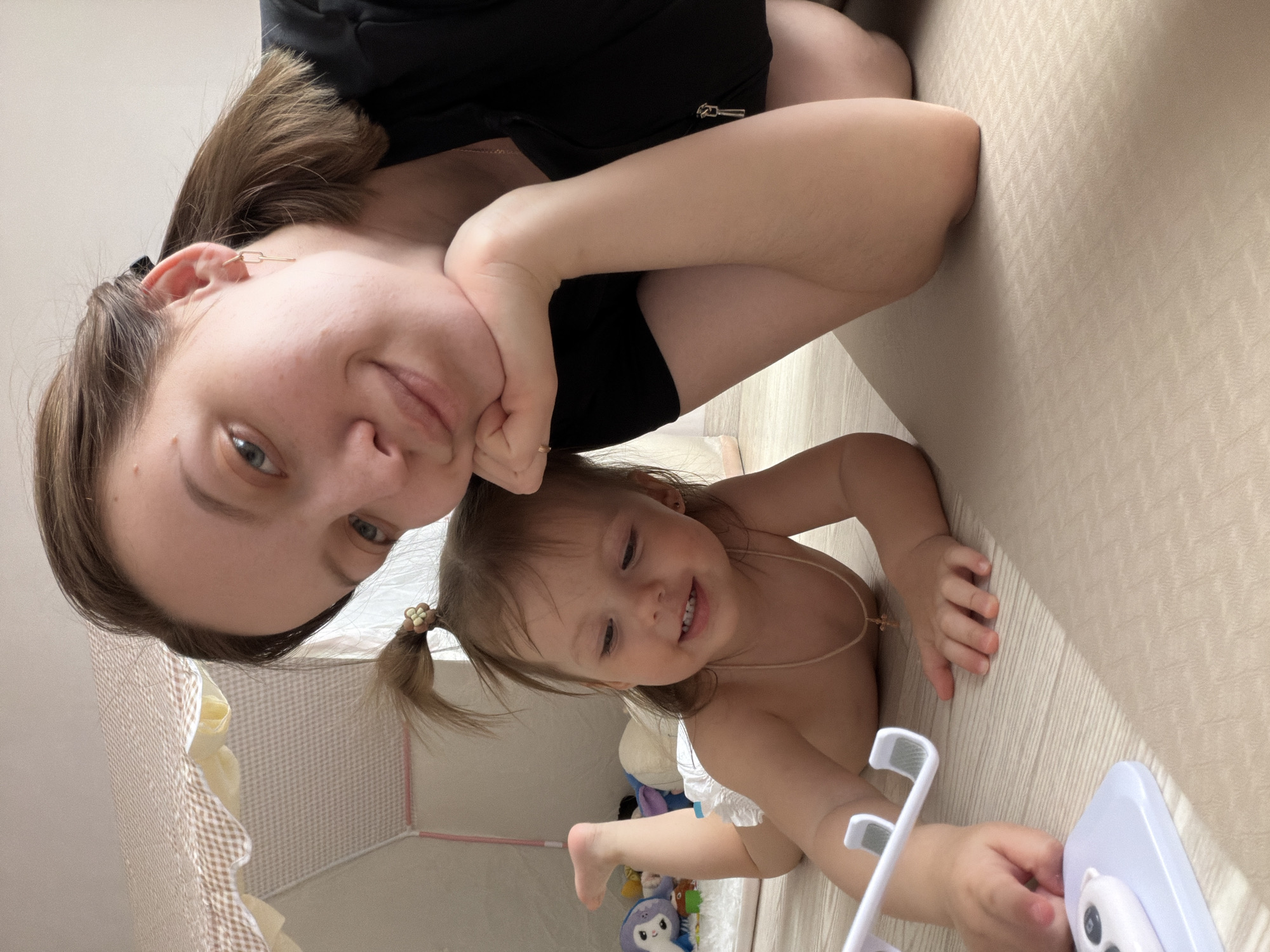 The image is a close-up, rotated photo of an adult female and a young child lying down indoors. The picture is rotated 90 degrees clockwise, presenting the subjects on their sides.
The adult, in the upper part of the rotated image, has light brown hair, fair skin, and blue eyes. She wears a black top and has her head resting on her right hand, looking directly at the camera with a gentle smile. A gold-linked earring is visible on her left ear, and a small gold ring adorns one of her fingers.
The young child is positioned in the lower right, below the adult. She has light brown hair tied in two small pigtails with flower-shaped hair ties and wears a white diaper and a thin necklace. The child is lying on her stomach, looking down and to her right with a wide, joyful smile that shows her teeth. Her right hand holds a white and light blue object, likely a toy, with white hook-like elements and small buttons. A blue and white plush toy is partially visible in the background near the child.
Both are on a light beige floor with a subtle textured pattern. In the background, a light-colored wall and a section of a playpen or bed with a checkered fabric liner are visible. The image captures a happy, casual moment between the adult and child in what appears to be a home setting in Reshetnikovo, Russia. Both subjects appear content and happy. There is no visible text.