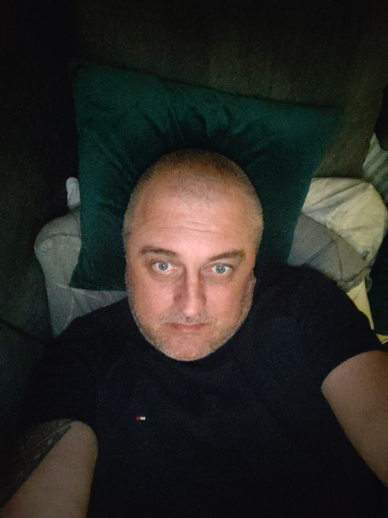 An adult male with light skin, closely cropped light-colored hair, and short facial stubble is depicted from a high-angle perspective, gazing directly into the camera. He wears a dark-colored, short-sleeved collared shirt that features a small rectangular red, white, and blue flag logo on the left chest. A tattoo is visible on his upper left arm. The man's head rests on a dark green pillow. To his left, a light-grey patterned cushion is present, and to his right, a striped light-colored fabric is visible. The surrounding environment is dimly lit and largely out of focus, indicating an indoor setting. This scene is located in Huntingdonshire, United Kingdom.