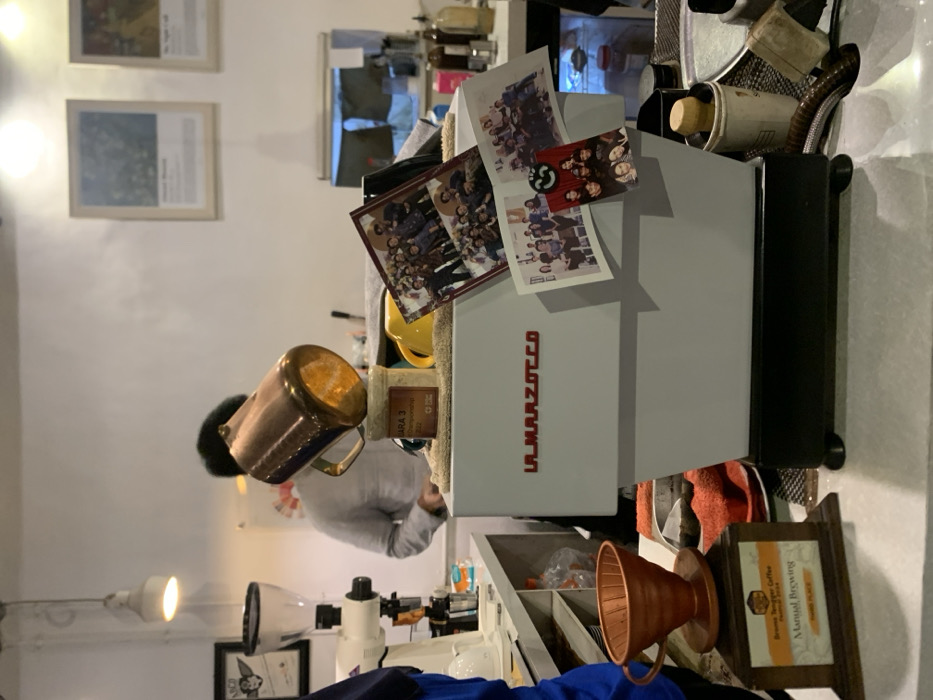 The image shows a coffee shop interior with a coffee machine, a barista, and various objects. The coffee machine is a La Marzocco, a brand of high-end espresso machines. The barista is standing behind the counter, wearing a gray shirt. A copper pitcher sits on the machine, and a jar labeled "BARA 3" with coffee beans is placed on the counter. There is a trophy next to the machine, with a black plaque that reads "Manual Brewing." There are also several photographs of groups of people attached to the coffee machine, presumably friends or coworkers. The image is taken in Probolinggo, Indonesia, specifically at a coffee shop located at -7.751 latitude and 113.213 longitude. The environment is a cozy and comfortable coffee shop with a modern interior.