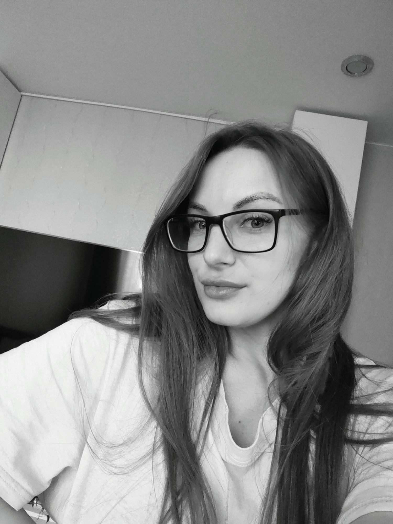 A black and white, close-up indoor portrait captures an adult woman from the chest up, with her gaze directed towards the viewer. She has long, dark hair that cascades over her shoulders and frames her face. She wears dark-rimmed rectangular glasses, which highlight her eyes, and a light-colored, casual t-shirt with a v-neckline. Her expression is calm and neutral, with a slight, subtle smile on her lips.
The background suggests an interior setting, possibly a home. To the left, there's what appears to be a section of an upper cabinet or storage unit, with a darker panel below it. A wall-ceiling corner with a thin molding runs horizontally across the top left. On the ceiling in the upper right, a recessed circular light fixture is visible. Another light-colored, rectangular vertical surface, possibly part of furniture or a door, is seen on the far right edge of the frame. The lighting appears even, suggesting an indoor light source. No visible text is present in the image. The monochromatic treatment emphasizes contours and textures, giving the image a classic feel.