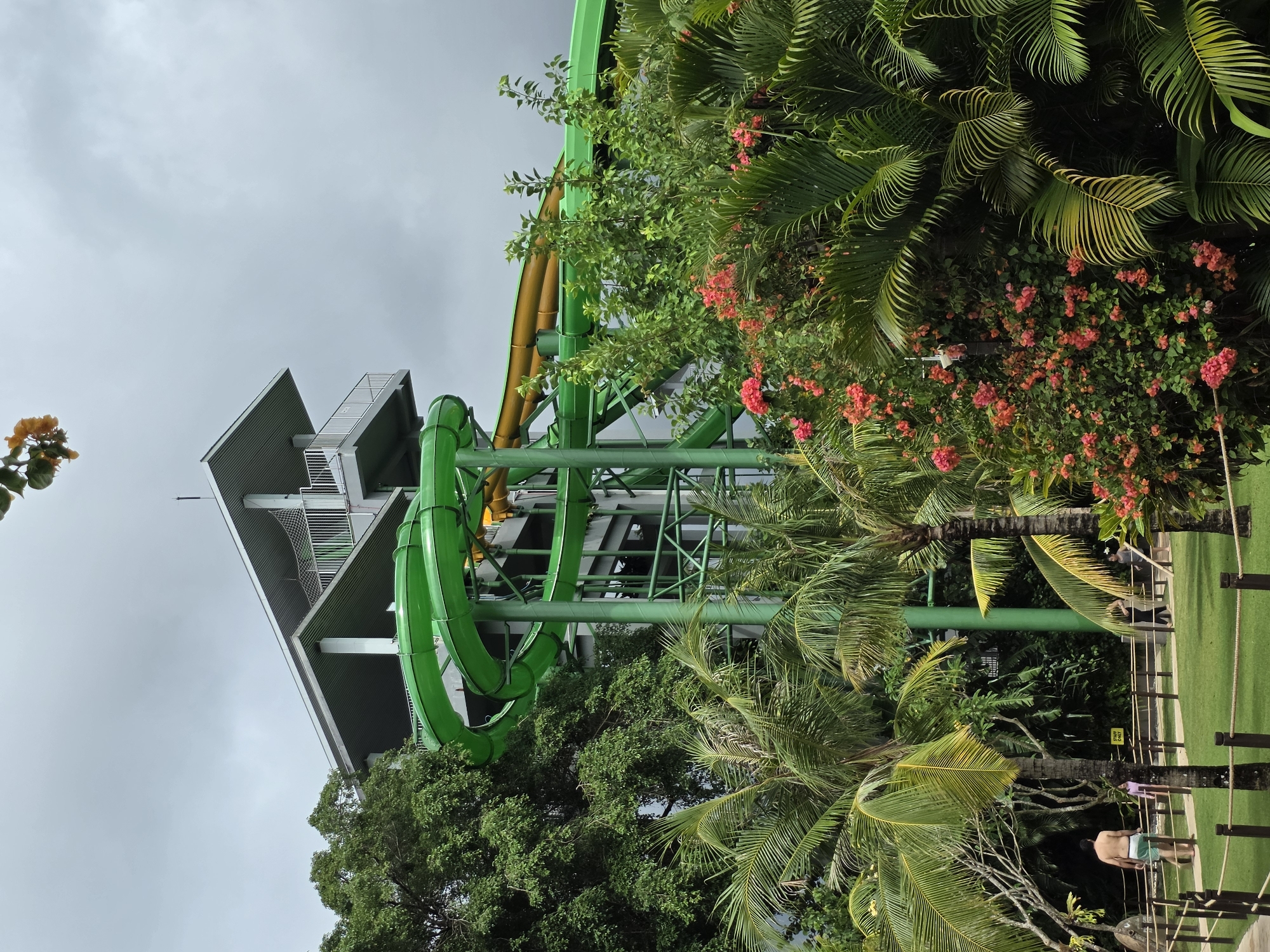 The image captures a water park scene in Kuta, Indonesia, likely Waterbom Bali. A large, multi-story structure with gray and white cladding supports several green and yellow water slides winding through the structure's framework. Dense tropical vegetation, including palm trees and flowering bushes with pink blossoms, surrounds the slides. The sky is overcast. In the foreground, a grassy area contains fence posts with rope and a few people. One man is shirtless, wearing green shorts, bending over. Another individual is visible near the fence.