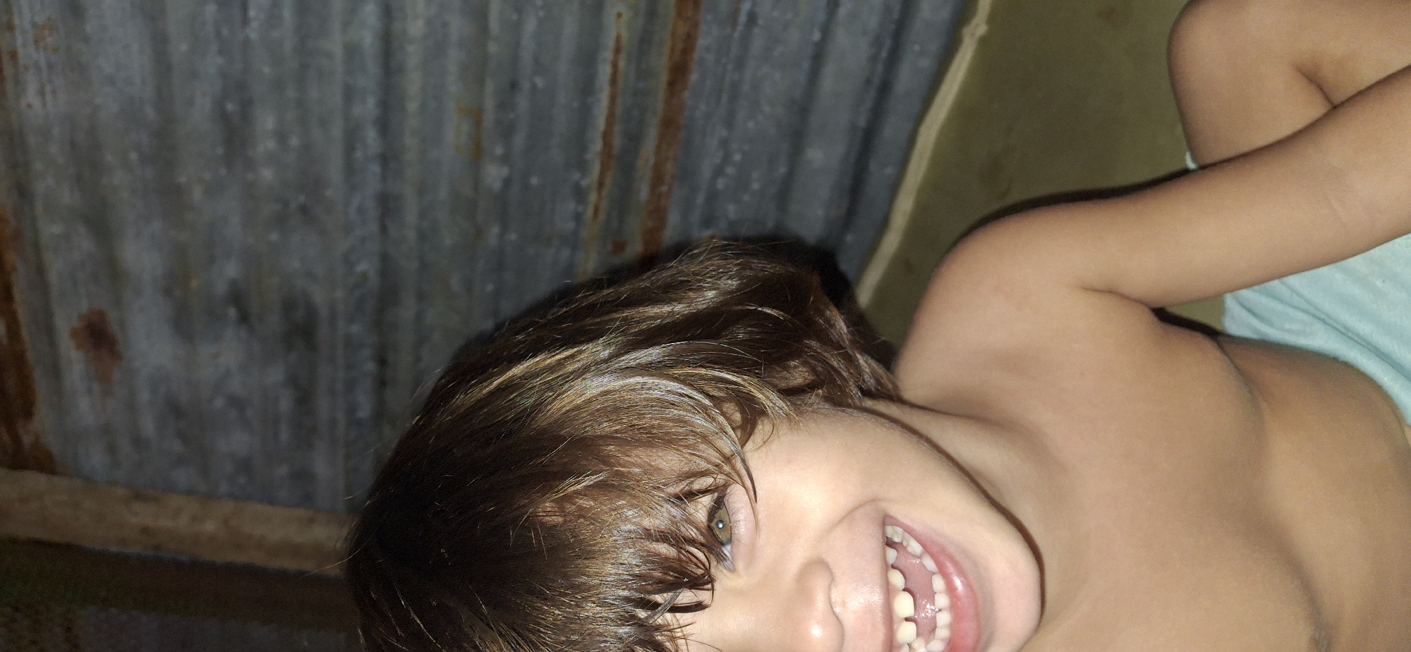 The image captures a close-up, diagonal view of a young child, likely a boy, against a rustic background in Vallecito, Venezuela. The child has light to medium brown, possibly wet or messy hair, and striking green or hazel eyes. He is smiling broadly with an open mouth, revealing his teeth, which conveys a strong sense of joy and playfulness. The child appears bare-chested, and a light blue garment is visible draped over his body on the right side of the frame.
The background is dominated by a weathered corrugated metal sheet, which exhibits signs of rust and age, suggesting an utilitarian or informal structure. A dark wooden beam runs horizontally across the lower left section of the background. Another wall or surface in a muted green-brown color is visible on the far right, behind the child's shoulder. The lighting is soft and diffused, illuminating the child's face and hair without harsh shadows, implying an indoor setting or a shaded outdoor area during the daytime. The overall impression is one of candid happiness in a simple, possibly rural or humble environment. No text is visible in the image.
