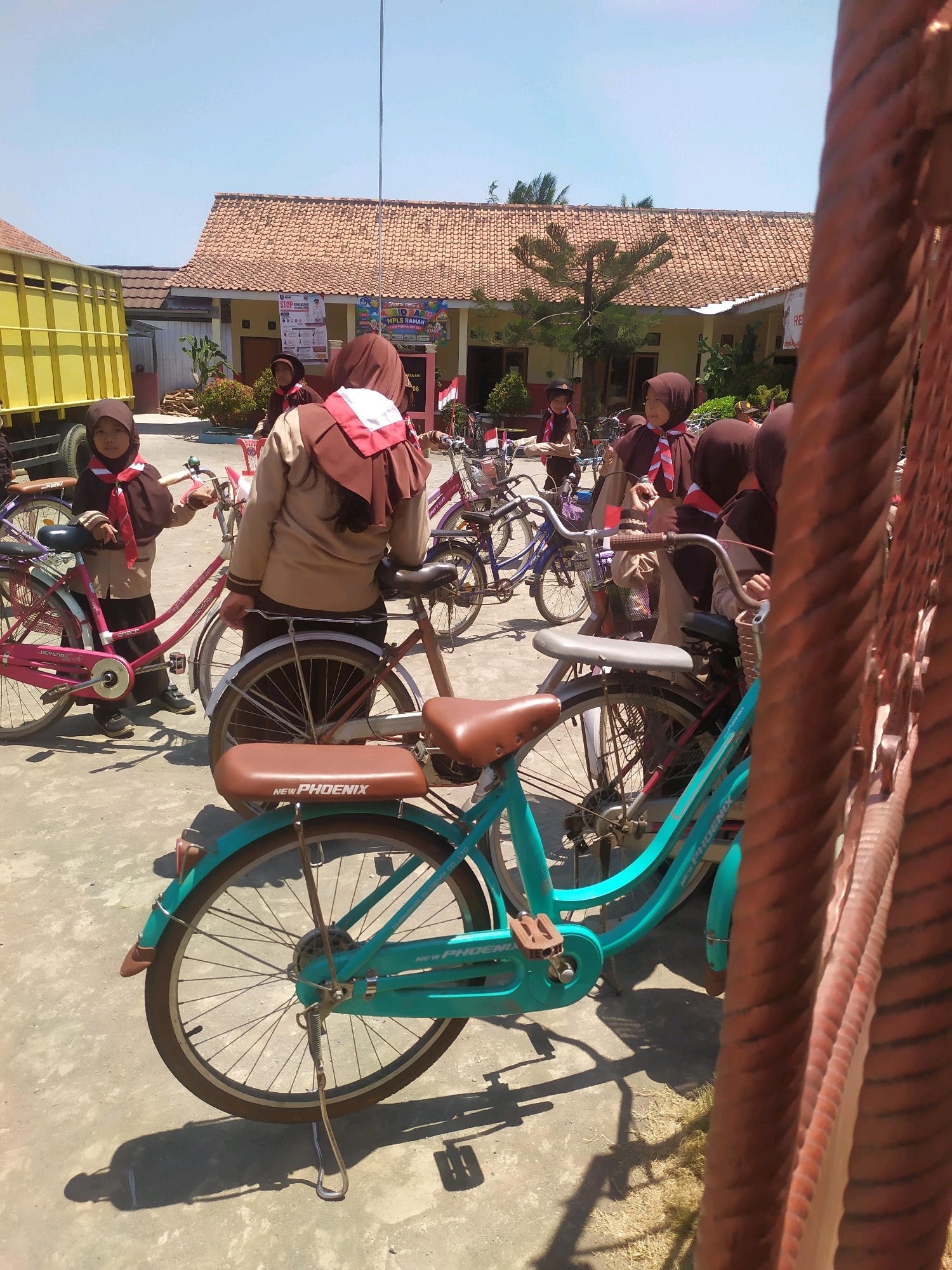 In an outdoor environment, a group of young girls in brown uniforms and headscarves stand next to several bicycles. The bicycles are of various colors, including pink and turquoise, and are parked on a paved surface. Behind the girls, a yellow truck is visible, and further in the background, a building with a tiled roof is present. The scene appears to be in a school or community area on a sunny day.