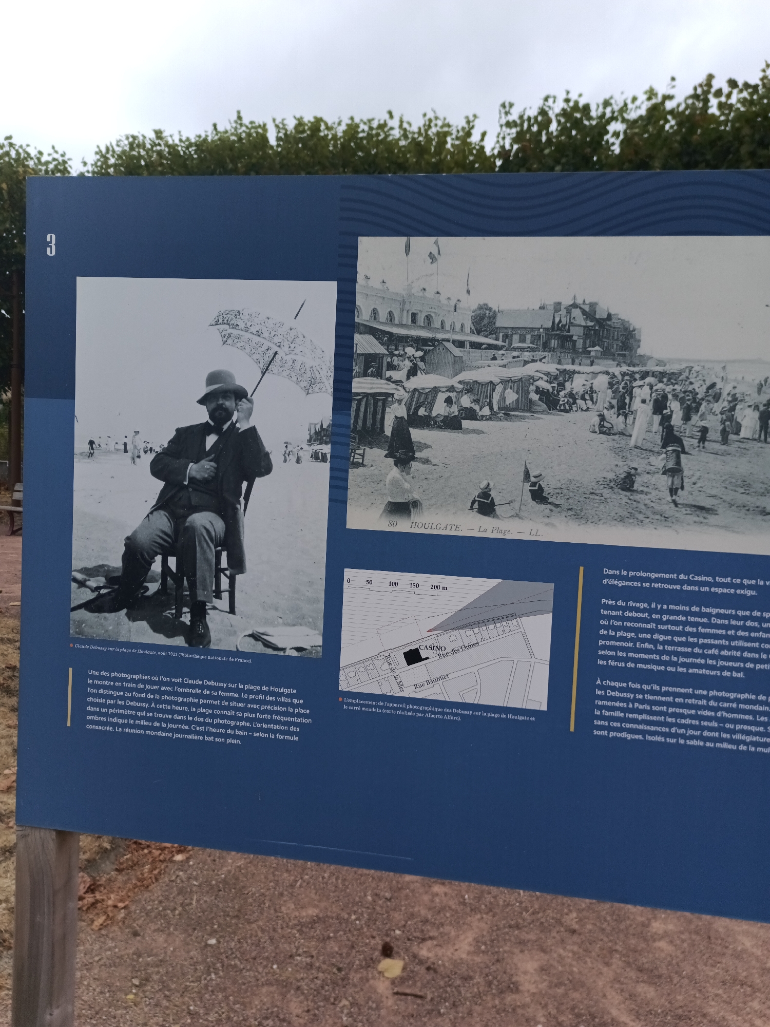 This is a photograph of an outdoor information display, likely in Houlgate, France. The display features a large blue background with white text and two historical black and white photographs.
The photograph on the left shows a man, identified as Claude Debussy, sitting on a chair on a beach. He is wearing a suit and hat and holding an umbrella. He is looking upwards, possibly at the sky or at someone off-camera. The beach appears to be populated with other people in the background.
The photograph on the right depicts a bustling beach scene with numerous people, beach umbrellas, and buildings in the background, identified as Houlgate. There are also what appear to be beach tents or cabanas.
Below the photographs, there is text detailing the images and their context. The text mentions Claude Debussy on the beach of Houlgate in August 1911. It also describes the scene as a lively gathering of people.
Further down, there is a map or diagram of the area, showing streets labeled "Rue de la Mer," "Rue Baurnier," and "Rue des Dames," and a building marked "CASINO." There is also a scale indicating distances of 0, 50, 100, 150, and 200 meters.
The text also provides historical details about the promenade, casino, and the beachgoers in Houlgate. It describes the atmosphere as one of elegance and social activity, with people enjoying the beach, the casino, and the musical entertainment. The text further discusses how photographs of Debussy were taken on the promenade, emphasizing the social and leisure aspects of the scene.