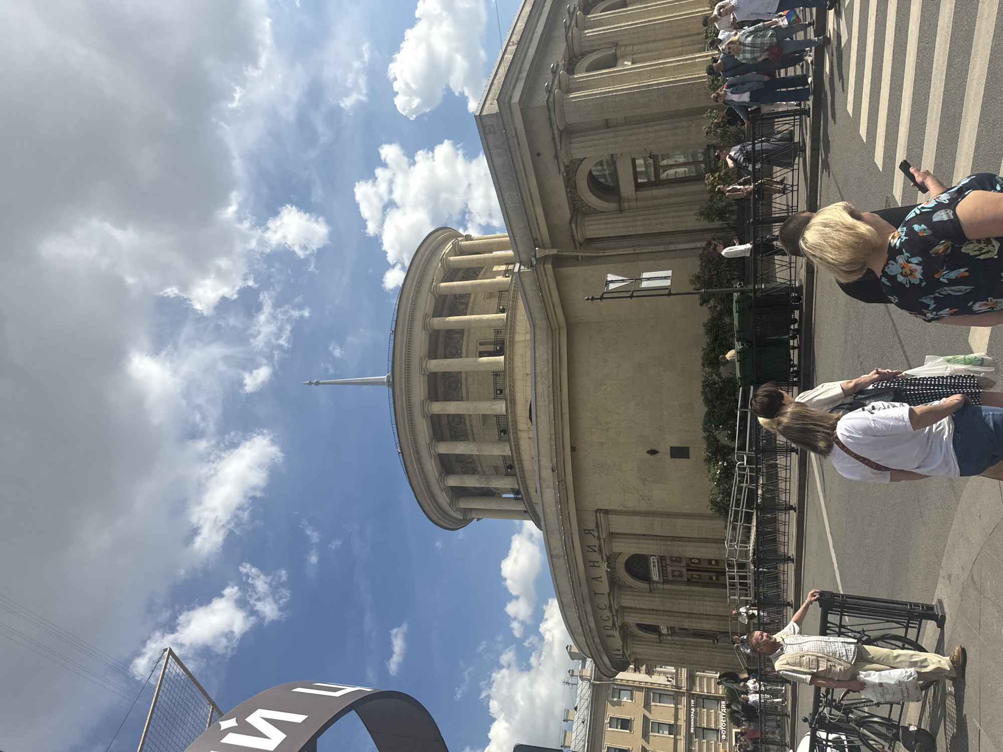 The image shows a wide-angle view of the exterior of the Moskovsky Railway Station (Moskovsky Vokzal) in Saint Petersburg, Russia, on a sunny day with scattered clouds. The station's distinctive rotunda-shaped upper level is visible, supported by a colonnade. The lower part of the building features arched windows and doors, and the word "ВОССТАНИЯ" (Vosstaniya) is inscribed on the facade, referencing the square it faces.
In the foreground, a street with a crosswalk is visible, with several people crossing or waiting. A man in a light-colored vest and checkered shirt stands near a black metal fence, holding a white plastic bag. To his right, two women are walking away from the camera, one in a white t-shirt and the other in a dark floral print top, both looking at their phones. Other pedestrians are seen in the middle ground, some walking along the sidewalk or near the building's entrance. Bicycles are parked to the left of the frame. The overall scene depicts a busy urban environment with a mix of architectural grandeur and everyday life.