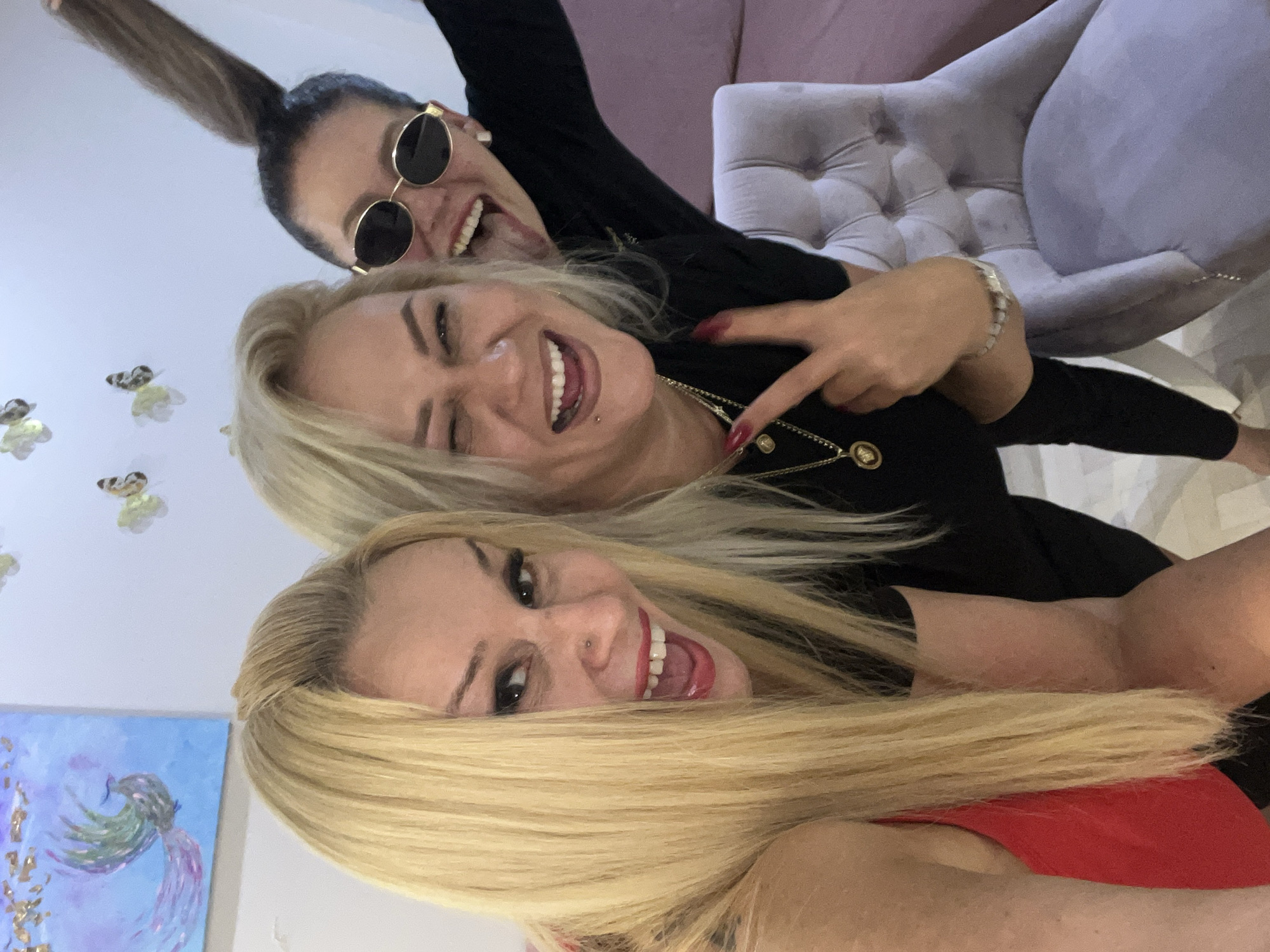 Three women are posing for a selfie in an indoor setting. The woman on the left has long blonde hair and is wearing a red top. The woman in the middle has blonde hair and is wearing a black top, with a piercing on her lip and a necklace. She is winking and making a peace sign with her left hand. The woman on the right has dark hair pulled up in a ponytail, is wearing sunglasses, and is sticking her tongue out. She is wearing a black top and earrings. In the background, there is a light blue painting of a bird with gold accents on the wall, and decorative butterflies. A grey velvet armchair is visible in the foreground. The location is not identifiable as Białystok, Poland.