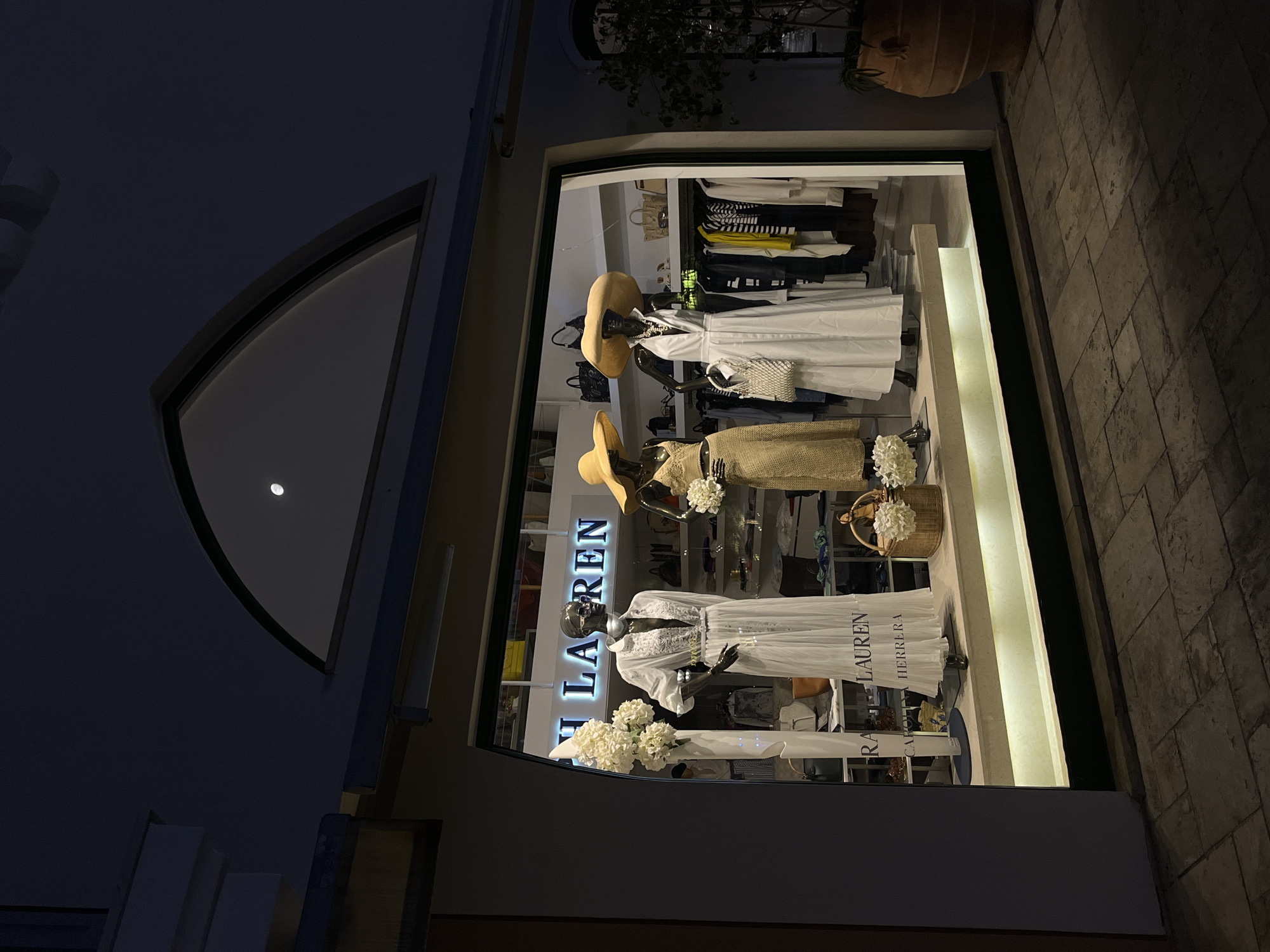In Limassol, Cyprus, at night, a brightly lit store window displays three mannequins wearing fashionable summer clothing. The store's name, "RALPH LAUREN," is illuminated above the display. The mannequins are adorned with large hats and dresses. To the left, one mannequin wears a long white dress, complemented by a bouquet of white flowers. Another mannequin in the center is dressed in a straw-colored dress and also holds flowers. The third mannequin on the right is styled in a white dress and carries a woven bag. The store window also reveals racks of clothing and accessories inside, including handbags, shirts, and dresses. The exterior of the store is dark, with the illuminated window creating a focal point against the night sky. The ground in front of the store is paved with stone tiles.