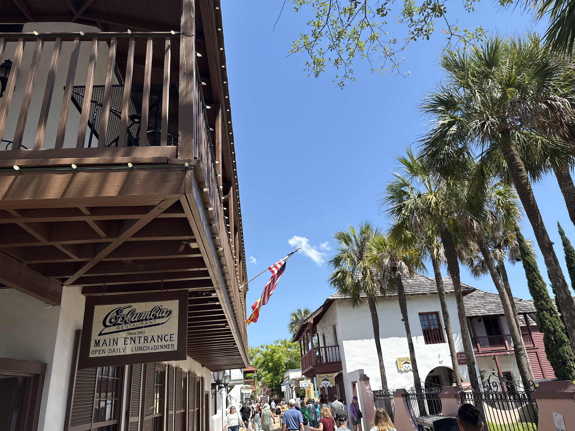 The image shows a street scene in Saint Augustine, Florida, United States. The left side of the frame features a portion of a building with a dark brown balcony and white facade. A sign reads "Columbia Restaurant, Main Entrance, Open Daily Lunch & Dinner." An American flag, flown with another, is visible suspended above the street.
The right side of the image displays palm trees lining the street and a white building with dark brown trim and a balcony. A sign with the address "97" is visible. People are seen walking on the street, a mix of locals and tourists. The sky is clear and blue with some tree branches and light green leaves are visible. The scene is well-lit by sunlight.