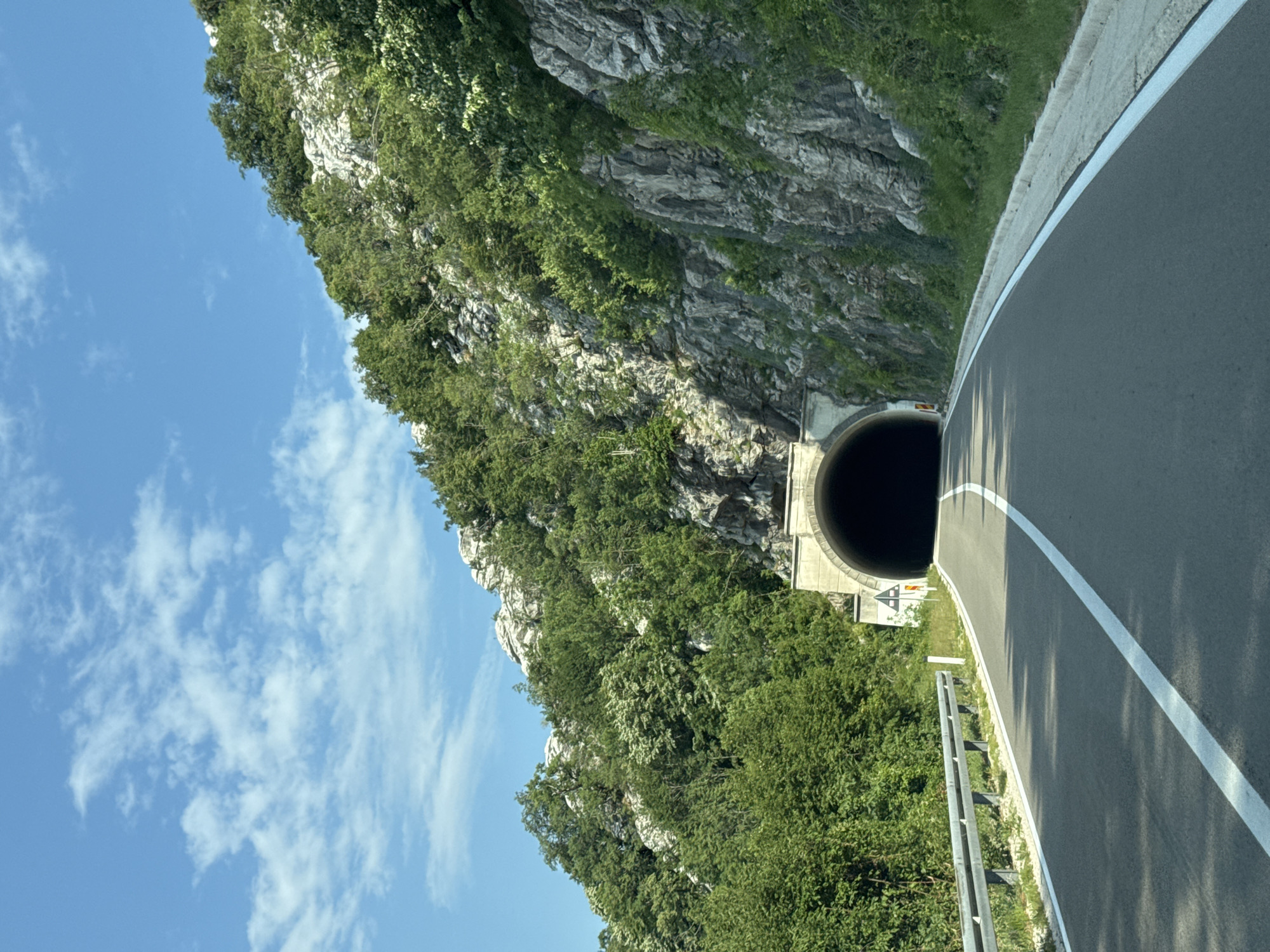 The image shows a view from a vehicle traveling on a paved road towards a tunnel entrance carved into a rocky, vegetated mountainside. The road has white lane markings. A metal guardrail runs along the left side of the road. The sky above is blue with scattered white clouds. The tunnel appears to be a concrete structure with a rounded archway leading into darkness. There are some traffic signs near the tunnel entrance. The surrounding environment is characterized by steep, rocky terrain covered in dense green trees and vegetation. This scene is located in the city of Dragalj, Montenegro.