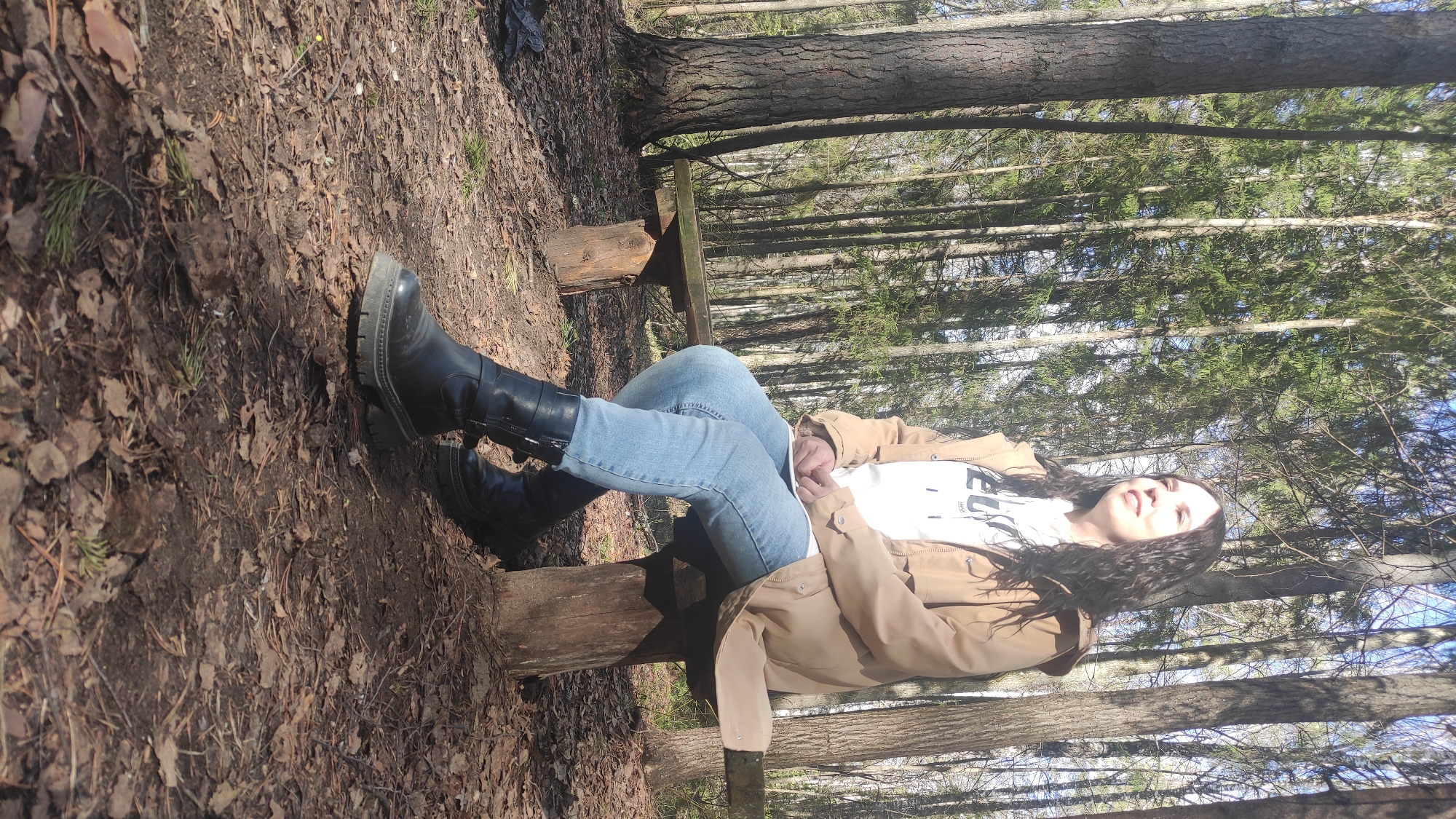The image depicts a woman sitting on a rustic wooden bench in a forest setting. The ground is covered with fallen leaves and scattered branches. The woman is wearing light blue jeans, black boots, a white t-shirt, and a light brown jacket. Her head is tilted upwards, and she is smiling. Behind her are numerous tall trees. The location is Unknown, Unknown.