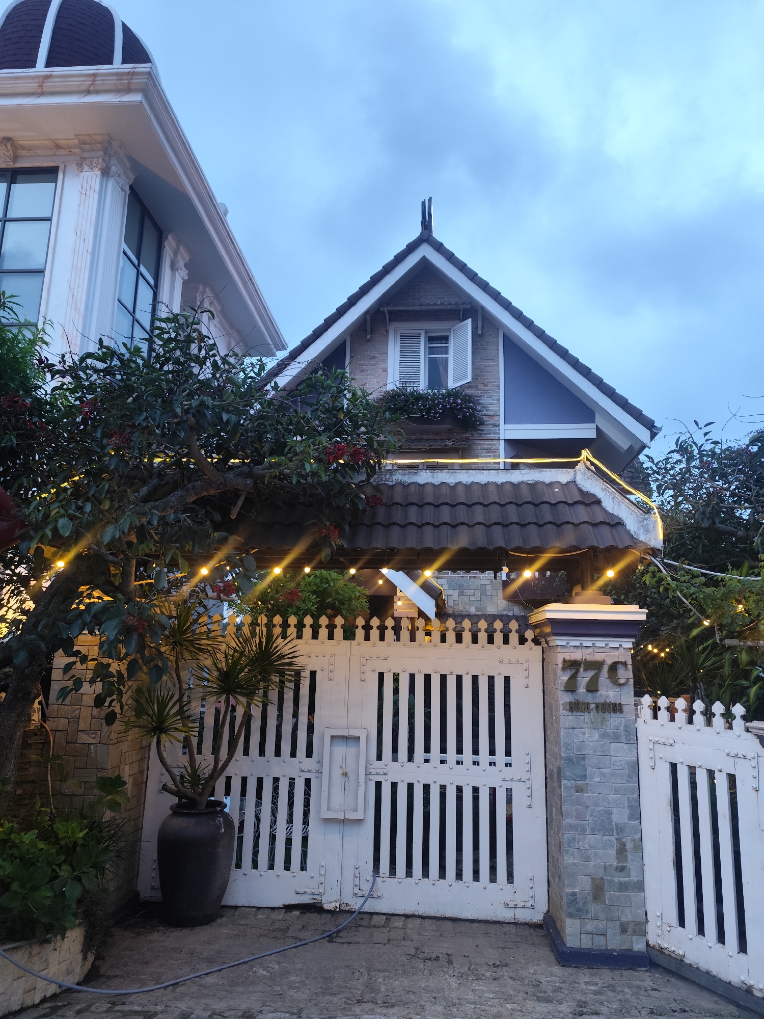 The image shows the exterior of a house with a white gate in Xuân Trường - Đà Lạt, Vietnam. The house has a gray tiled roof and a white facade. A tree with red flowers is in front of the house, decorated with string lights. A potted plant sits next to the gate, which has the house number "77C" on a stone pillar. The sky is overcast and it appears to be dusk. There are no people visible in the image.
