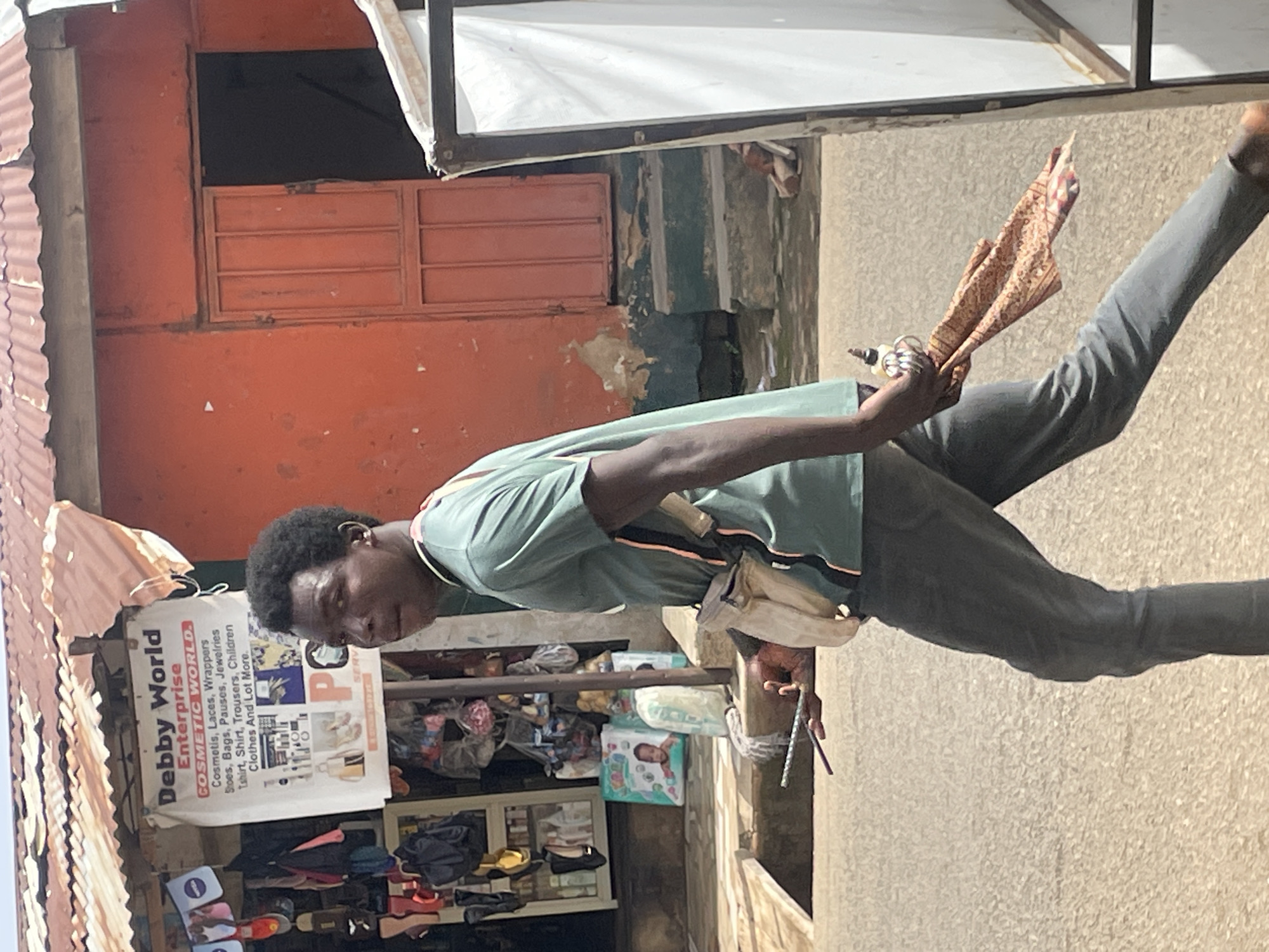 A young man, possibly of Nigerian descent, is captured mid-stride on a sun-drenched street, his gaze directed off-camera towards the right. He is clad in a casual, short-sleeved green t-shirt with subtle pink and black trim along the collar and sleeves, paired with dark grey trousers. A light-colored satchel is slung across his chest, resting on his hip. In his right hand, he clutches a patterned cloth, possibly a scarf or wrap, and a small, dark object. His left hand holds a few thin, dark rods.
The background reveals a bustling urban scene, likely in Gura Top, Nigeria. A weathered, corrugated metal roof shelters a storefront adorned with a white sign that reads "Debby World Enterprise" and lists various goods, including "COSMETIC WORLD. Cosmetis, Laces, Wrappers, Shoes, Bags, Pauses, Jewelries, Tshirt, Shirt, Trousers, Children Clothes And Lot More." To the right of the sign, a vibrant orange-painted wall with a metal door is visible, suggesting a commercial establishment. Further to the right, a white structure with a dark frame stands, potentially a shop display or billboard. The street itself is paved with what appears to be gravel or small stones. The overall impression is one of everyday life in a Nigerian marketplace.